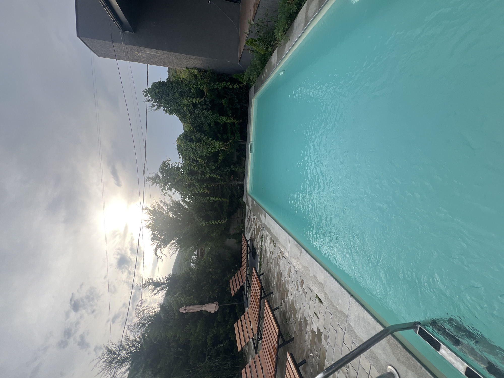 The image shows a swimming pool in Tsodoreti, Georgia. The pool is filled with clear turquoise water and is surrounded by a paved patio. There are several wooden lounge chairs arranged on the patio, and a large umbrella is also present. Lush green trees and vegetation line the perimeter of the area, with a modern dark-colored building visible on the right. The sky above is cloudy with the sun peeking through, casting a soft light on the scene. A metal ladder provides access to the pool. The overall impression is one of a serene and relaxing outdoor space.