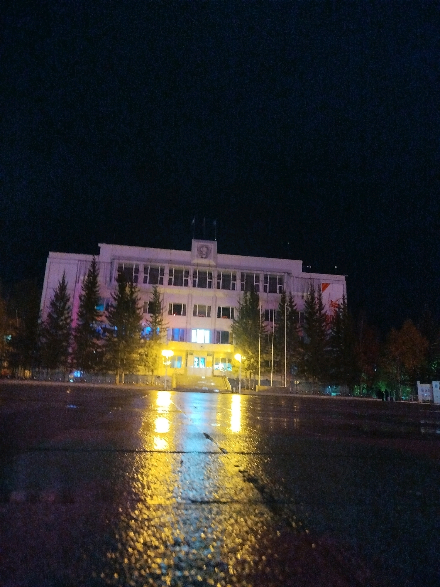 This is a nighttime, low-angle shot of the Palace of Culture in Mirny, Russia, after a rainfall. The wet asphalt in the foreground reflects the bright yellow and blue lights from the building and street lamps. A multi-story building with a light-colored facade is visible, featuring numerous windows, some of which are illuminated. Several tall evergreen trees stand in front of the building, and to the right, there are deciduous trees with some autumn foliage. The sky is dark and clear, typical of a night sky.