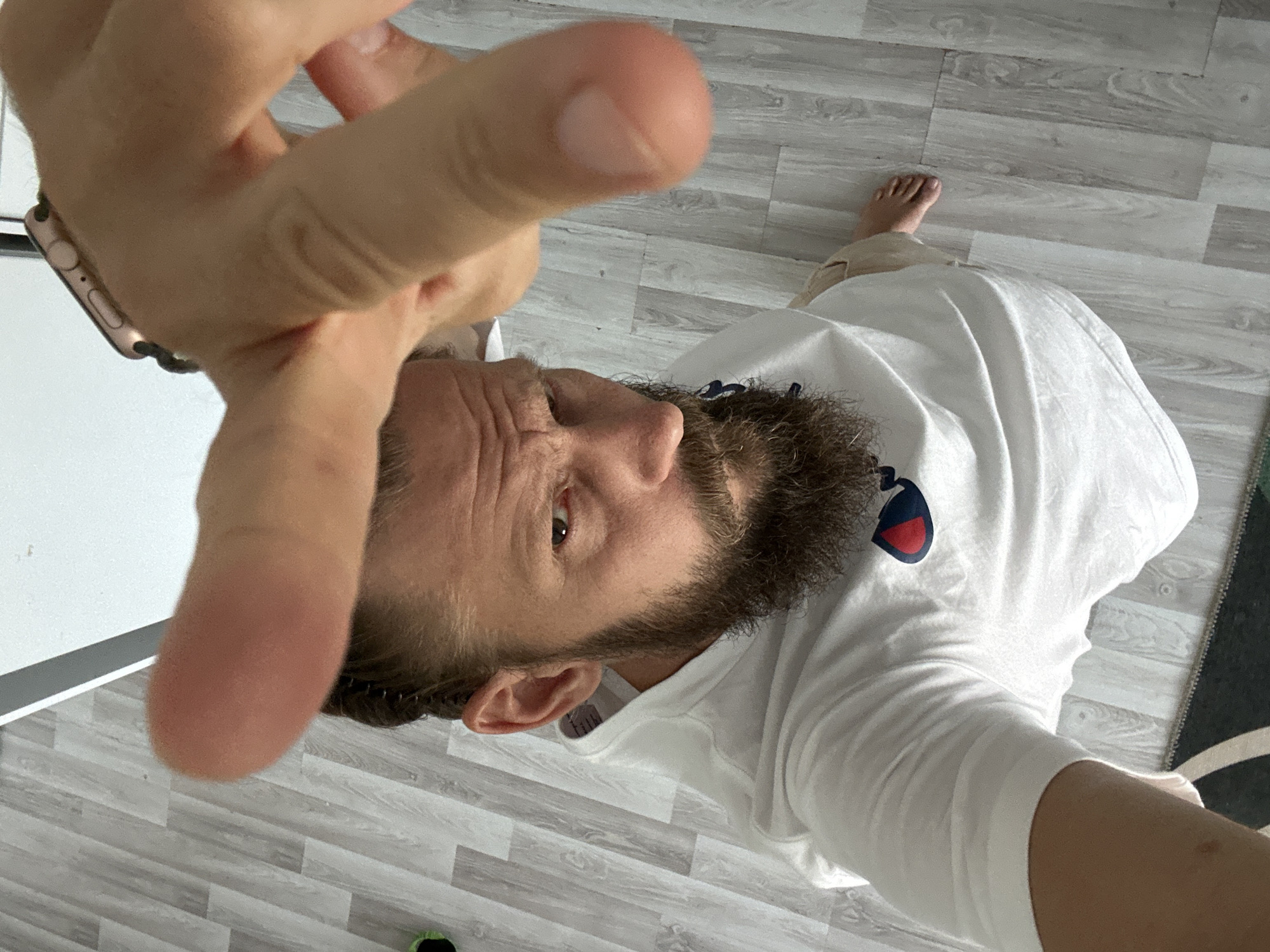 A man with a beard and a white t-shirt is looking up at the camera, with his hand in the foreground. The background appears to be a wooden floor. The man is wearing a watch on his left wrist. This image was not taken in Fethiye, Turkey.