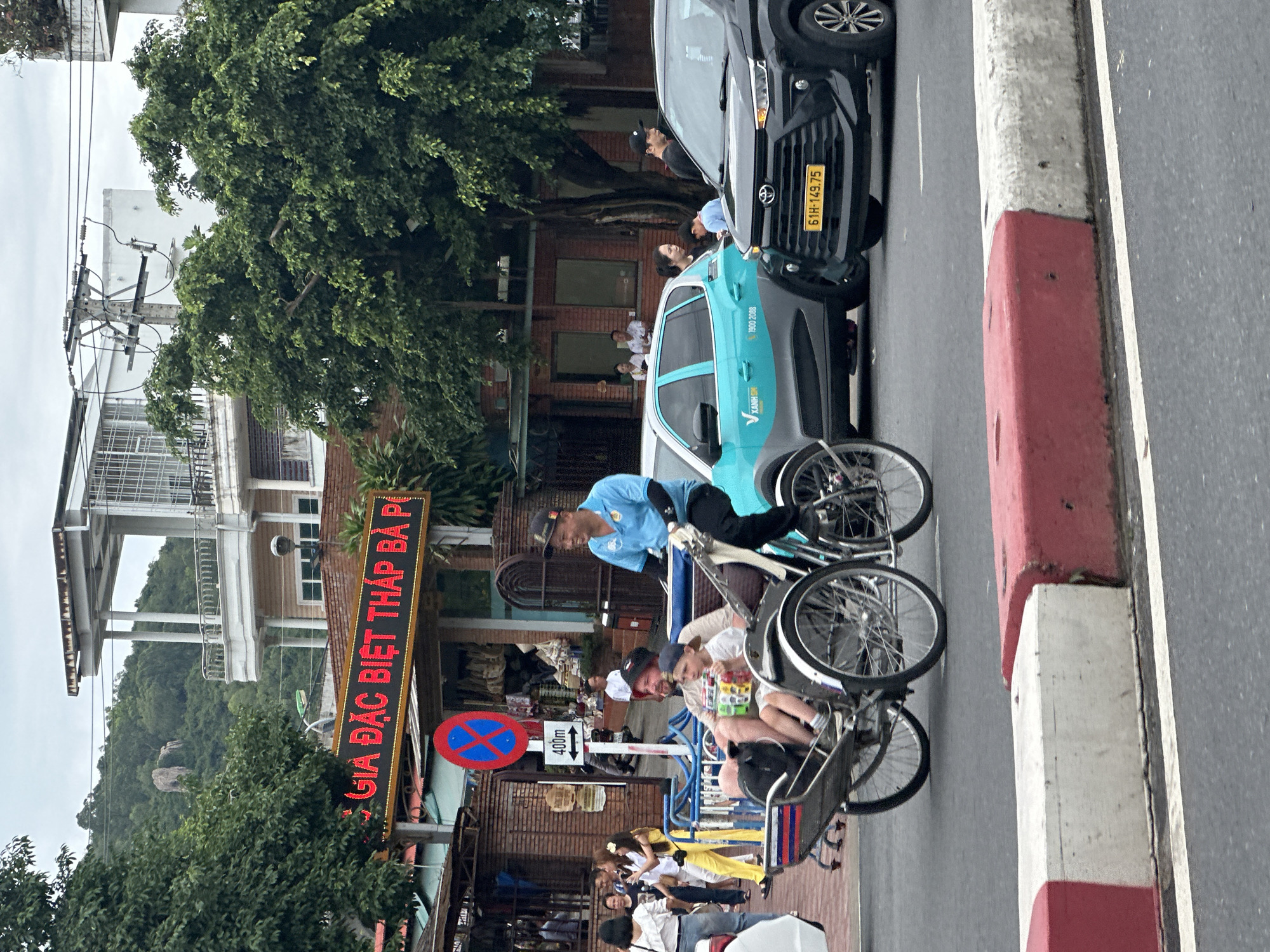The image shows a street scene in Bac Nha Trang Ward, Vietnam. A cyclo, a bicycle-powered three-wheeled vehicle, is in the foreground. The driver, wearing a blue shirt and cap, is pedaling. Two passengers, including a child, are seated in the front. Behind the cyclo are parked cars, one black and one light blue. People are standing near the cars. In the background is a building with a red neon sign that says "GIA ĐẶC BIỆT THÁP BÀ P" (likely a restaurant or business name) and a "No Entry" sign. Other people are visible seated near the building. The curb is painted red and white. The weather appears overcast.