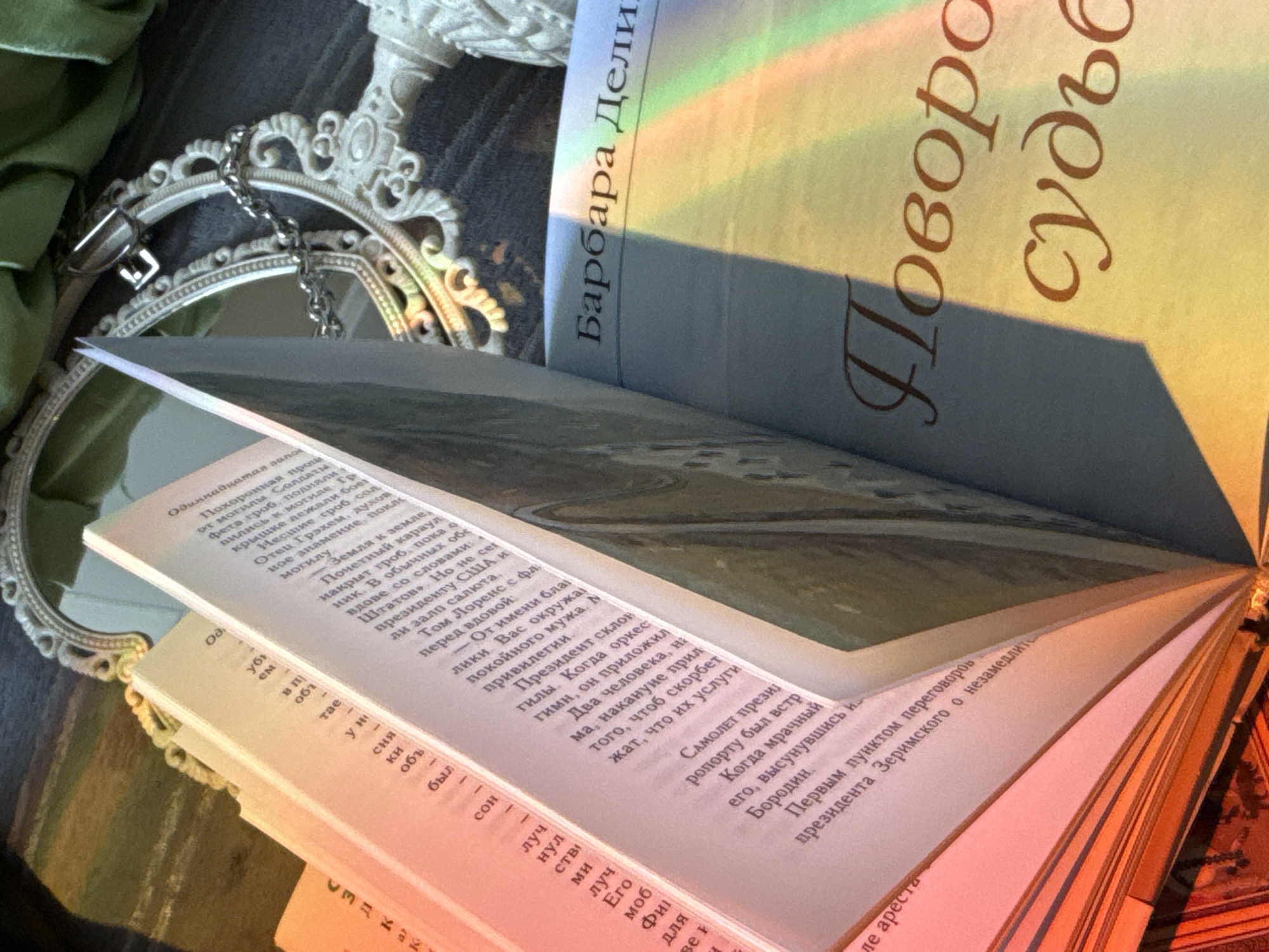 The image displays a still life arrangement centered around books and a decorative mirror, illuminated by a prominent rainbow light. An open book with white pages filled with Cyrillic text rests partially on an ornate, white-framed mirror. A silver-colored chain with a lock or clasp is draped over the mirror's frame.
Behind the open book, another book stands upright, its cover reflecting a vibrant rainbow spectrum. The author's name, "Барбара Дели" (Barbara Deli), and the title, "Поворот судьбы" (Turn of Fate or Twist of Destiny), are visible in Cyrillic script on the cover. A snippet of green fabric is visible on the left side of the frame, and a partial view of an ornate, white decorative object, possibly a candle holder, appears in the upper right.
The open pages of the book reveal several readable phrases in Cyrillic, including "Одиннадцатая авлов" (Eleventh of August), "Похоронная прош" (Funeral procession), "Могилы Солдати" (Graves of Soldiers), "президента США" (US President), "От имени бла" (On behalf of), "Президент склон" (President is inclined), "САМОЛЕТ Презид" (President's plane), and "Первым пунктом переговоров президента Зеримского о незамедлит" (The first point of negotiations of President Zerimsky about immediate...). These details suggest the book might be a novel with political or historical themes.
The rainbow light, likely from sunlight passing through a prism or window, indicates a daytime setting, casting a serene and artistic glow over the scene. The objects are arranged to create a thoughtful and aesthetically pleasing composition.