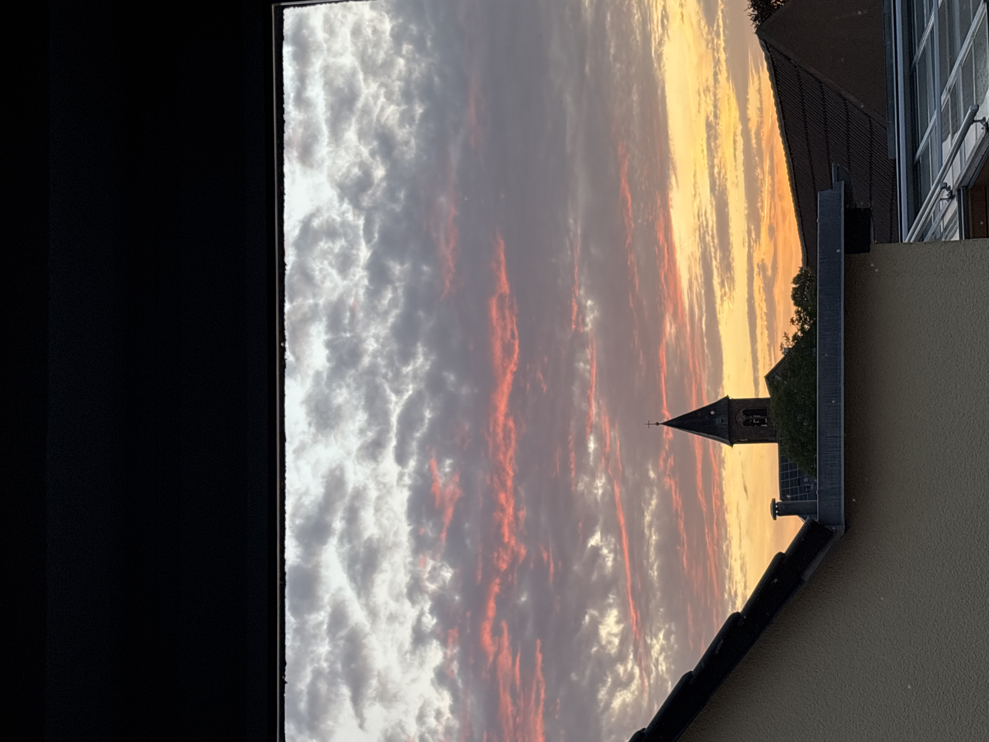 This image captures a sunset viewed through an opening, likely a window or a framed aperture. The sky is filled with clouds that are illuminated by the setting sun, displaying hues of pink, orange, and gray. In the mid-ground, a church tower with a cross on top rises above a cluster of trees and a building with solar panels. The foreground is dominated by the rooftops of several buildings, one on the left with a cream-colored facade and dark tiled roof, and another on the right with a dark tiled roof. The image appears to be taken from an elevated position, looking outwards at the sky and the urban landscape.
Specifically, this scene is located in Durmersheim, Germany, as indicated by the presence of the specific church tower and the architectural style of the buildings visible. The overall atmosphere is serene, emphasizing the natural beauty of the sunset against the backdrop of the town. There are no people visible in the image.