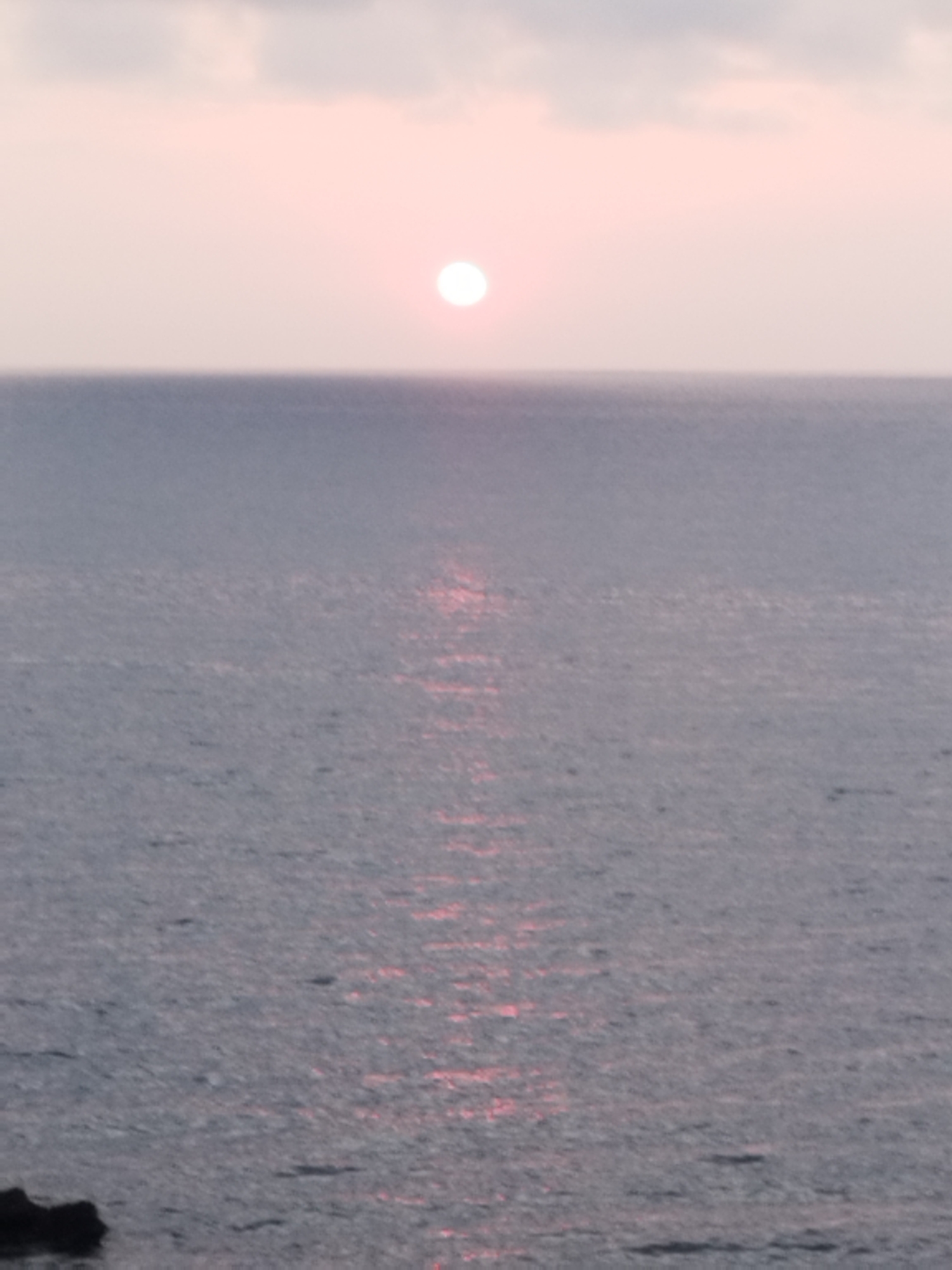 The image depicts a serene seascape with the sun low on the horizon, casting a soft pink glow across the sky and water. The sun is a bright, circular orb positioned slightly left of the center, with a shimmering reflection extending down the water's surface. The sea is a muted gray-blue, with gentle ripples visible. In the lower left corner, a dark, jagged rock formation protrudes slightly from the water. The overall impression is one of peaceful twilight. While the image is a beautiful depiction of a coastal scene, it does not contain any identifiable elements that would allow for its specific location within the city of Antibes, France.