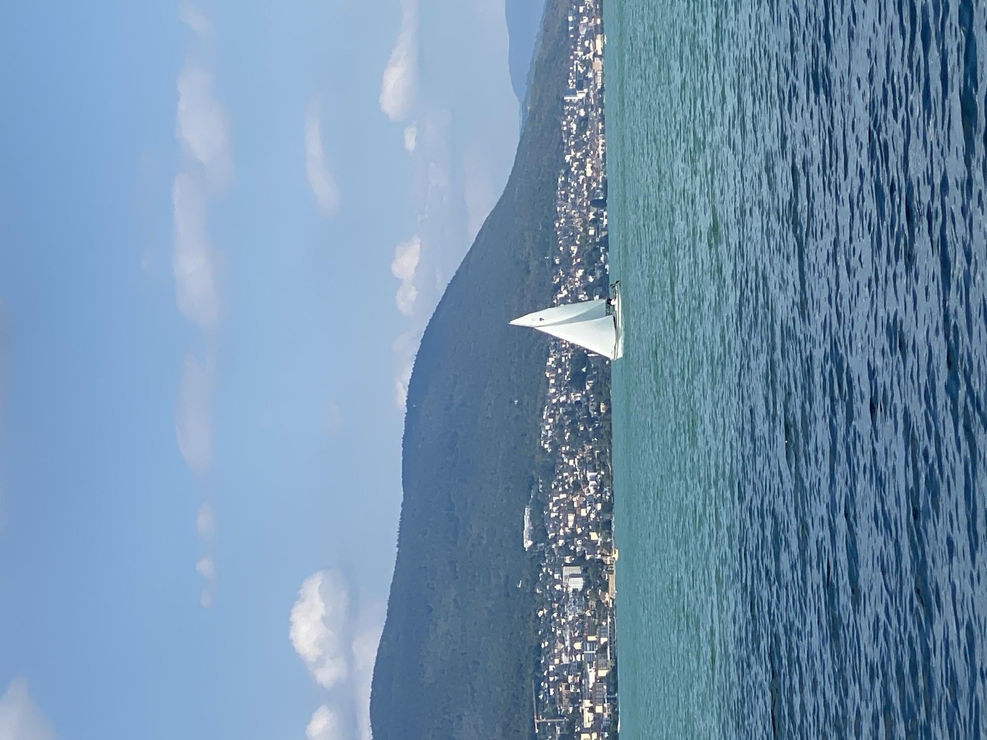 A sailboat with a white triangular sail navigates a body of water under a partly cloudy sky. The sailboat is positioned to the right of the frame, with a small crew visible on board. In the background, a densely populated shoreline city stretches horizontally across the image, backed by a large, tree-covered mountain. The water in the foreground is a darker blue, transitioning to a lighter turquoise hue closer to the shore. The city appears to be Cortaillod, Switzerland, situated on the banks of Lake Neuchâtel. The scene is tranquil and evokes a sense of leisure.