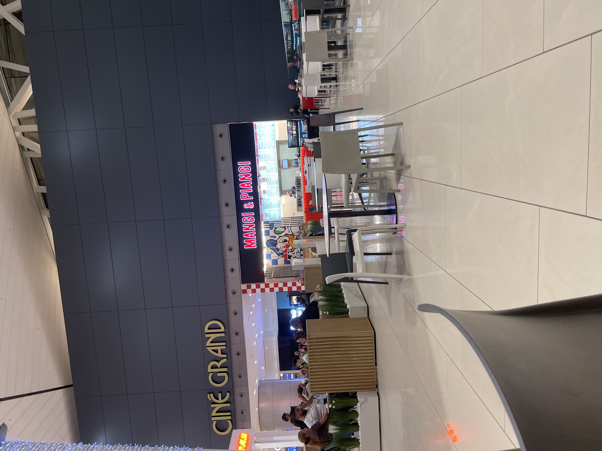 The image captures a scene in a shopping mall, likely located in Sofia, Bulgaria.
The building features signage for "CINE GRAND" in large gold letters and "MANGI & PIANGI" in red and white text, indicating a cinema and a food establishment within the mall. The architecture is modern, with grey tiled walls.
Several people are present, some appearing to be dining at tables associated with "MANGI & PIANGI". There are rows of white chairs and tables.
The floor is light-colored tile. The mall's interior includes modern design elements. The lighting is bright, suggesting an open and inviting environment.