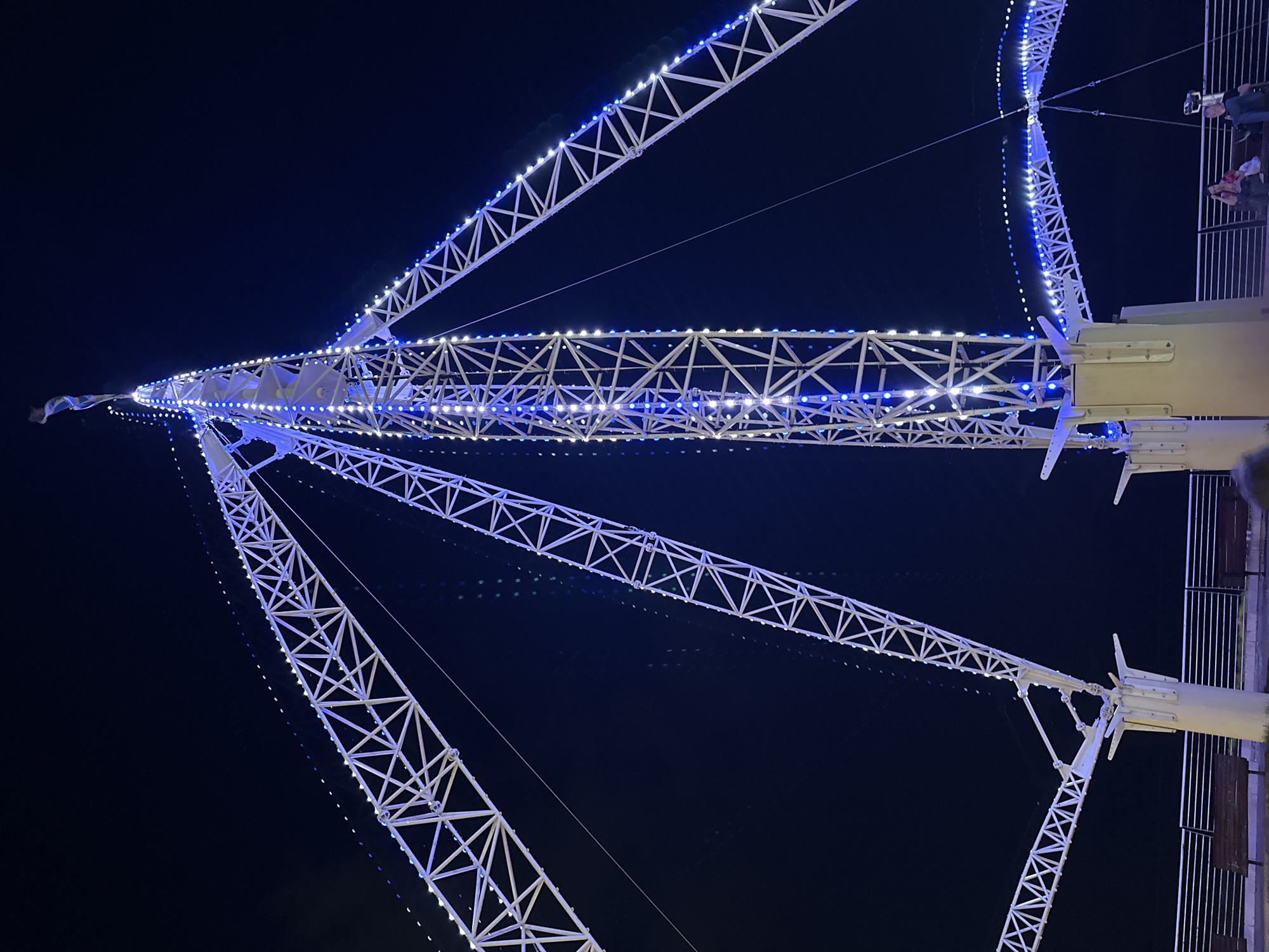 The image depicts the "Sky Drop" attraction at Luna Park in Nahariyya, Israel, at night. The attraction is a tall, white, lattice-like tower structure illuminated with blue and white lights. Several angled arms extend from the central tower, also adorned with lights. In the foreground, there are benches and railings, suggesting a public area. Two people are visible sitting on a bench in the lower right corner of the frame, one of whom is looking at their phone. The sky is dark, indicating it is nighttime, and the overall scene is brightly lit by the attraction's lights.