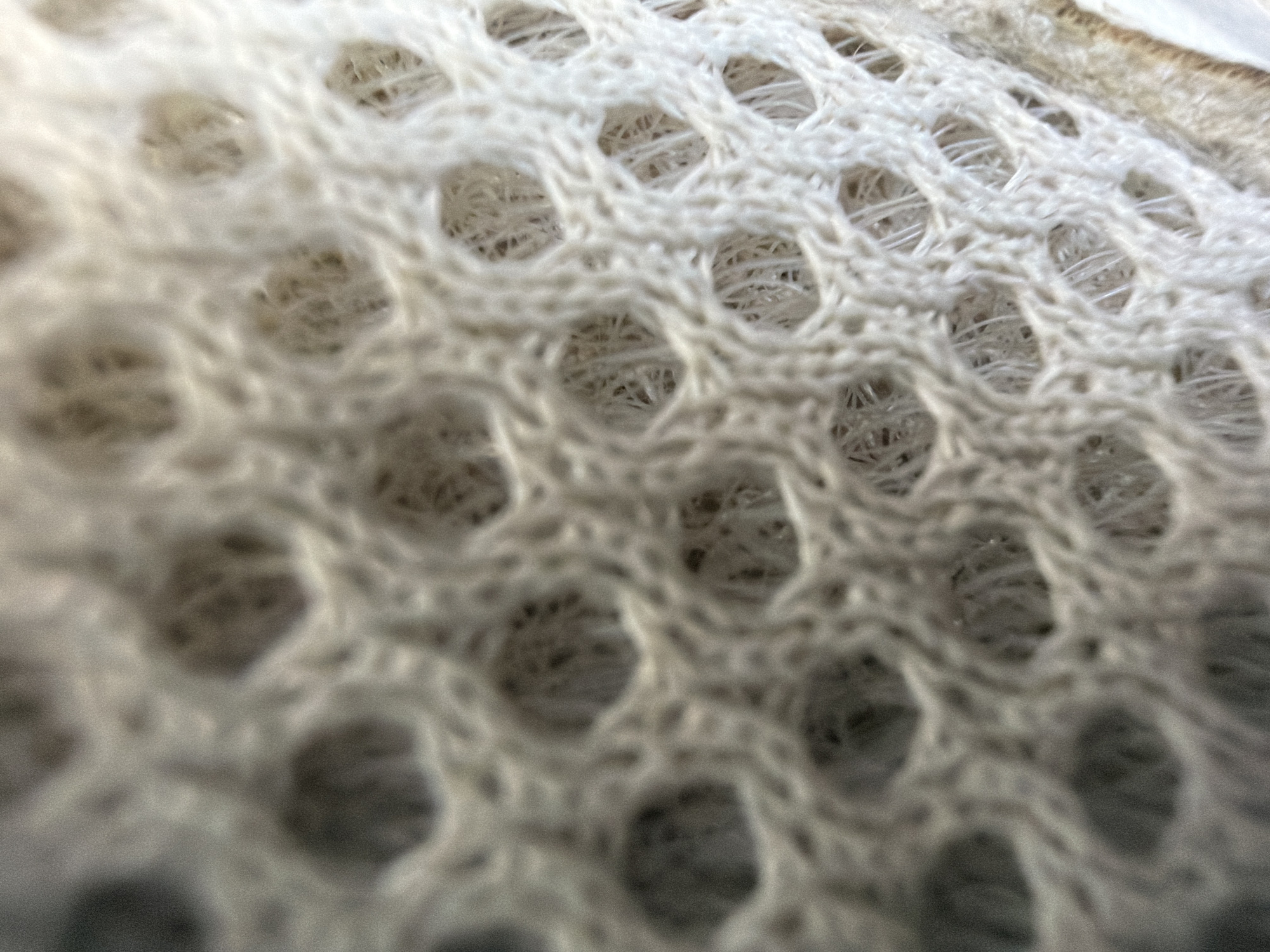 This image is a close-up, macro shot of a white, woven fabric with a honeycomb pattern. The texture is soft and slightly fuzzy, with visible threads forming a grid of hexagonal openings. The lighting creates subtle shadows within the weave, highlighting the three-dimensional structure of the material. There are no people, objects, or any indication of a city location, including Minsk, Belarus. The image focuses solely on the intricate details of the fabric's texture and pattern.