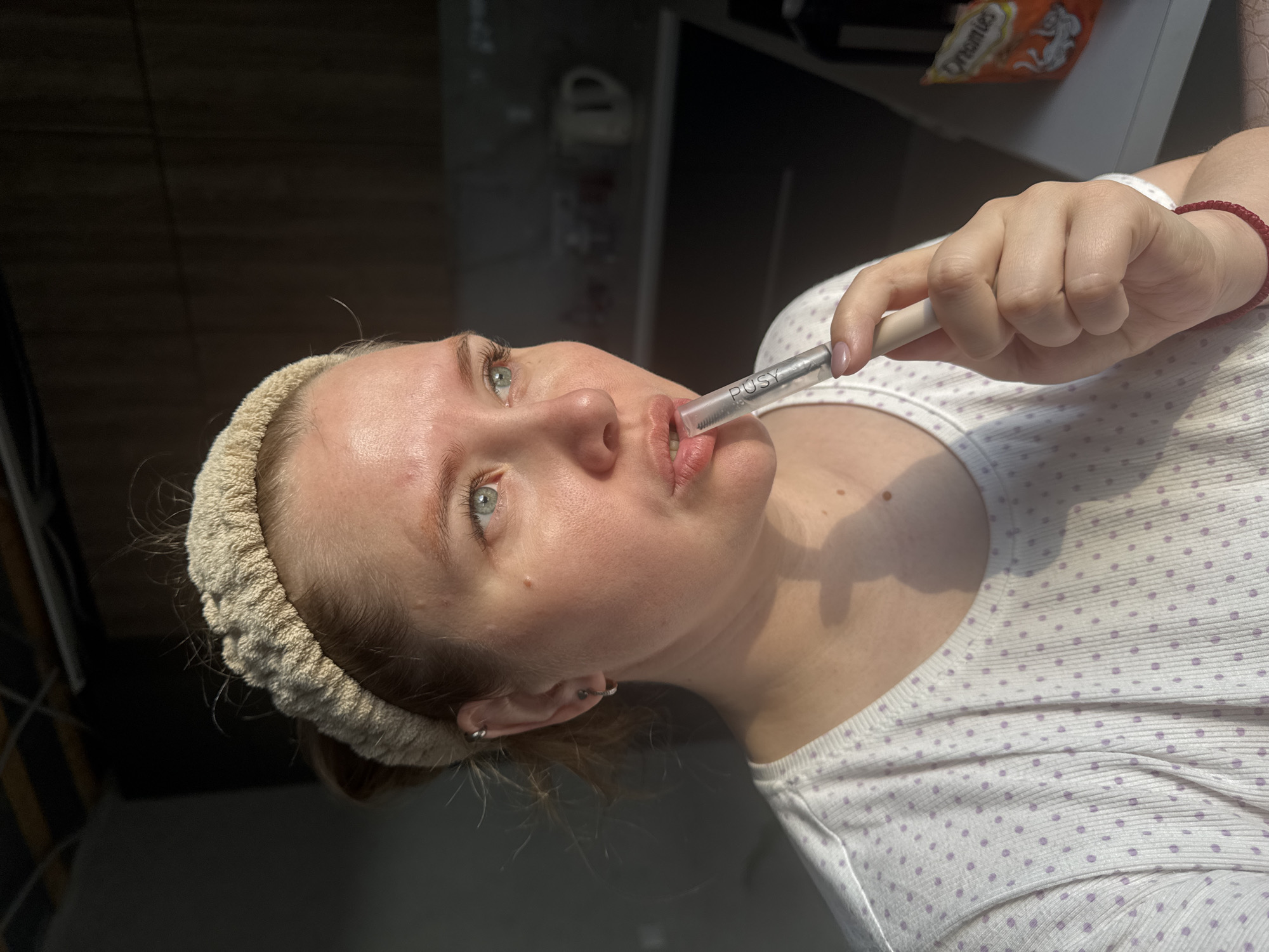 A woman with fair skin, light hair, and blue eyes is holding a clear tube with a white top in her mouth. The word "PUSY" is visible on the tube. She is wearing a white top with purple dots and a beige headband. She has multiple earrings in her left ear and a red beaded bracelet on her right wrist. The background appears to be a kitchen or bathroom area. The city of Ферма, Russia is not readily discernible based on this image alone.