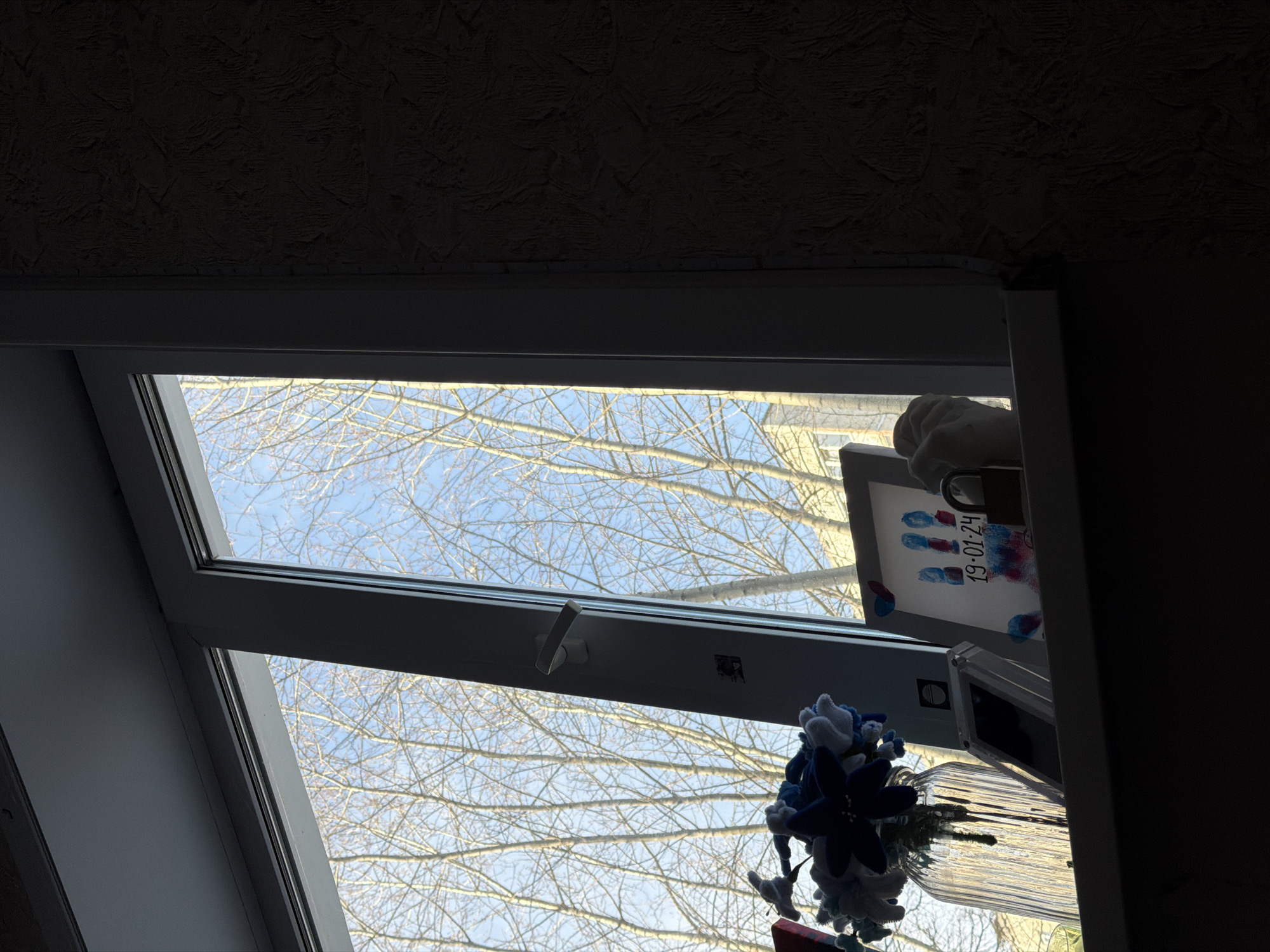 The photograph shows an interior view through a roof window, likely in Perm, Russia, given the Cyrillic characters on a card stating "19-01-24". The window reveals leafless branches against a pale blue sky. A vase with blue and white artificial flowers sits on the sill. A framed artwork depicting blue and red handprints and the date "19-01-24" leans against the window, secured by a padlock. The room is dimly lit, with dark textured ceilings visible. The window frame is gray. A portion of a light-colored sculpted hand is visible near the frame, obscuring a portion of the artwork.
