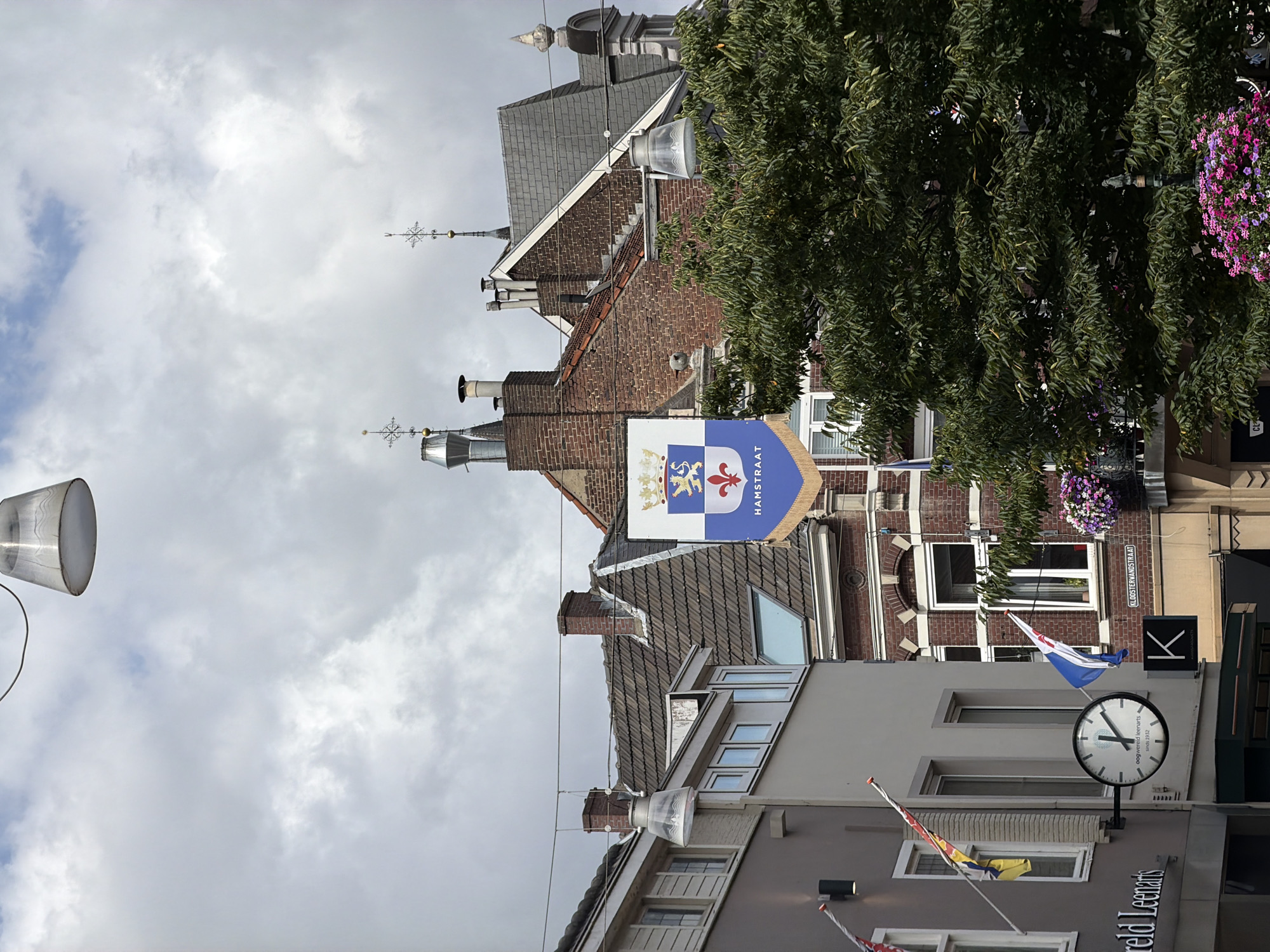 The image depicts a street scene in Roermond, Netherlands, under a cloudy sky. A banner with a coat of arms, reading "HAMSTRAAT," hangs from a wire strung across the street. The banner features a blue and white shield with a lion and a fleur-de-lis.
Buildings line the street, characterized by their tiled roofs, brickwork, and various architectural styles. Several chimneys are visible on the rooftops. On the left side of the image, a light-colored building has windows and flags attached to its facade. A round clock is mounted on this building, displaying the time as approximately 10:07. Below the clock, text reads "OOGVERBLIND Leenarts SINCE 1932."
To the right of the clock, a building with red brick and white accents features multiple windows. Another banner, with red and white stripes, is attached to this building. A street sign reading "KLOOSTERWANDSTRAAT" is visible on this building.
In the foreground, a large green tree with dense foliage dominates the right side of the frame. Below the tree, colorful flowers, predominantly pink and purple, are displayed in hanging baskets. A street lamp is visible amidst the foliage. The overall impression is of a European street with historical architecture and a vibrant display of nature.