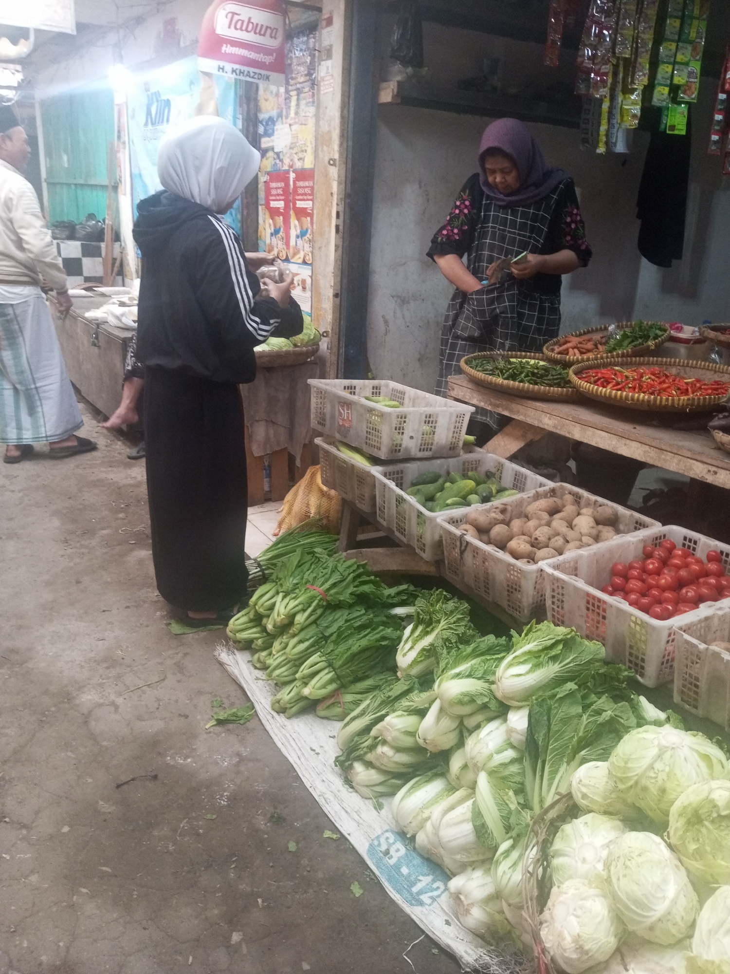 This image depicts a street market scene in Kalibeber, Indonesia. A woman wearing a purple hijab and a checkered dress is standing behind a stall, holding a small piece of paper and what appears to be a small object in her hands. In front of her are woven baskets filled with various vegetables, including red chilies, green beans, and what look like carrots. Another person, wearing a white head covering and a black hoodie with white stripes, stands facing away from the camera, looking at the stall's offerings and holding a clear plastic bag with some items inside. To the left, another individual, partially visible, is dressed in a white sarong-like garment and a light-colored top. The market stall is stocked with fresh produce, including large bunches of leafy green vegetables, cabbages, and tomatoes displayed in white plastic crates. In the background, there are shelves with packaged goods and signs, one of which prominently displays the word "Tabura." The ground is paved with concrete.