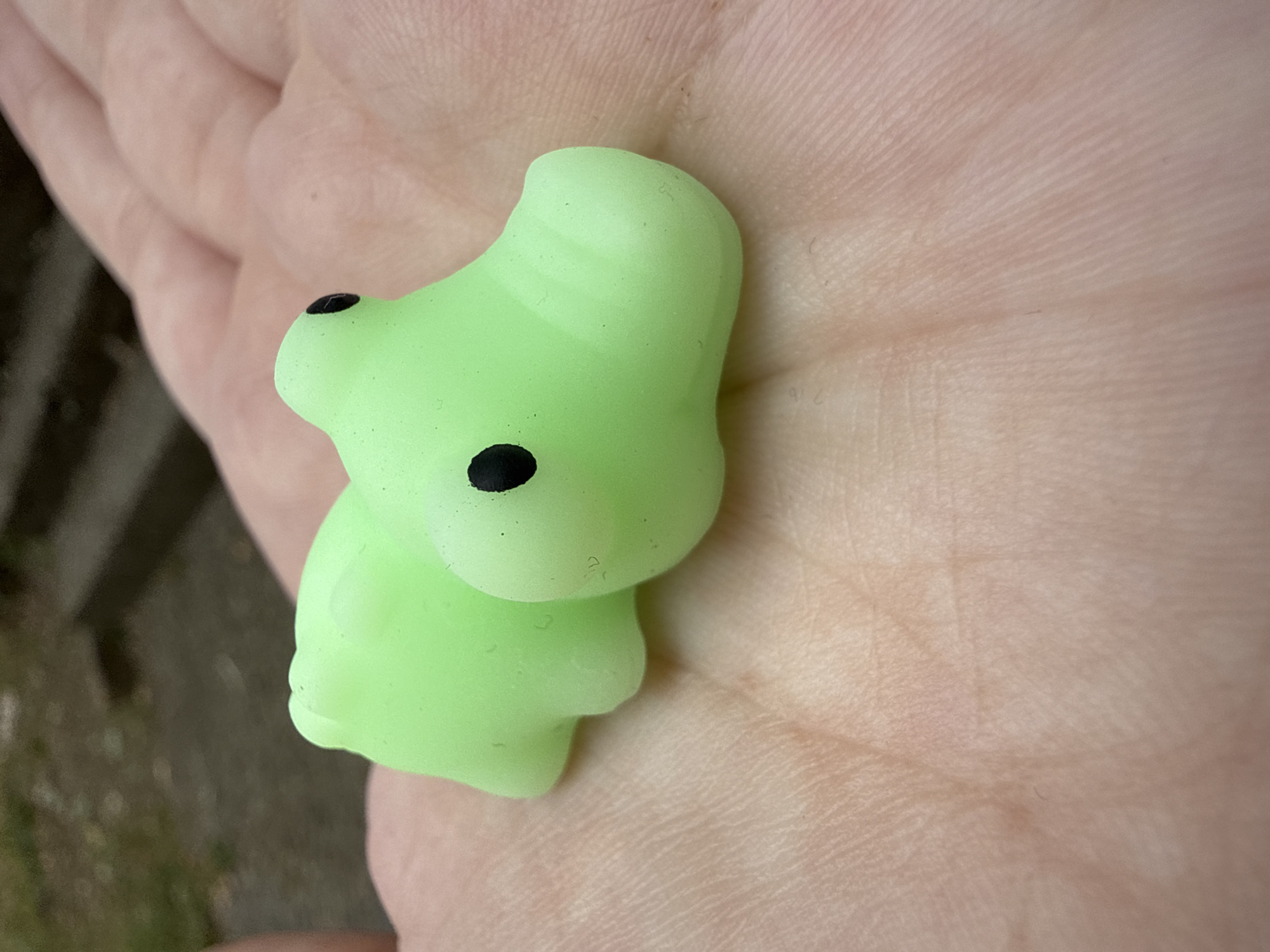 A pale green, alligator-shaped stress toy rests on the palm of a person's hand. The toy is smooth and appears to be made of silicone. It has two black dots for eyes and a rounded snout. The hand is lightly tanned with visible lines and creases. In the background, out of focus, is a blurry outdoor scene with hints of greenery and possibly stairs or a pathway. The scene is consistent with a casual, everyday moment. There is no discernible location in Burgas, Bulgaria indicated in the image.