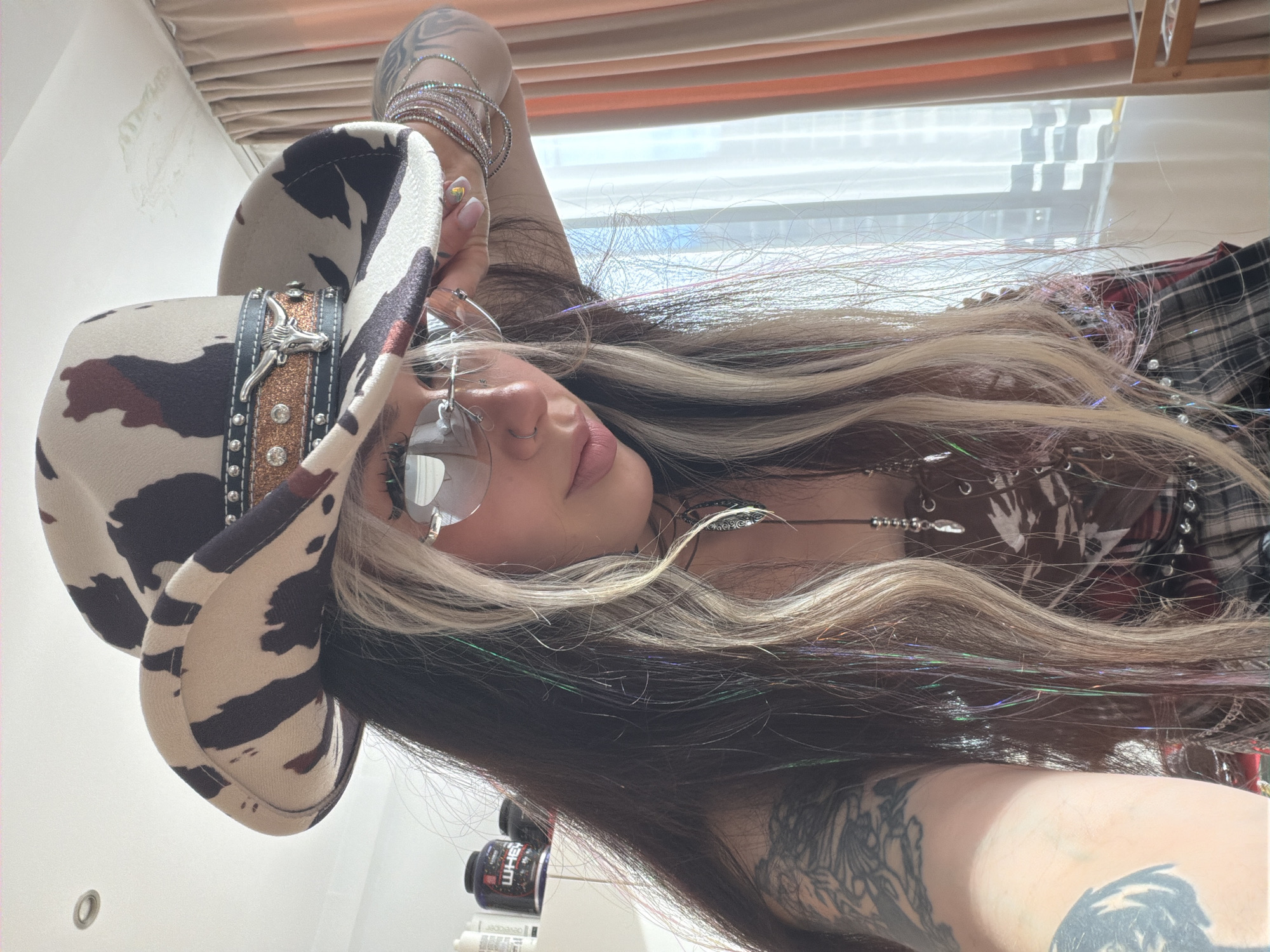 A woman is posing for a selfie. She is wearing a cow-print cowboy hat adorned with a bull skull and rhinestones. She also wears wire-rimmed glasses, a necklace, and numerous bracelets on her arm. Her hair is dark with blonde streaks and tinsel strands. Tattoos are visible on her arm and hand. The background shows a window with blinds and bottles/containers on a shelf. The location and city of Budva, Montenegro cannot be determined from the image.