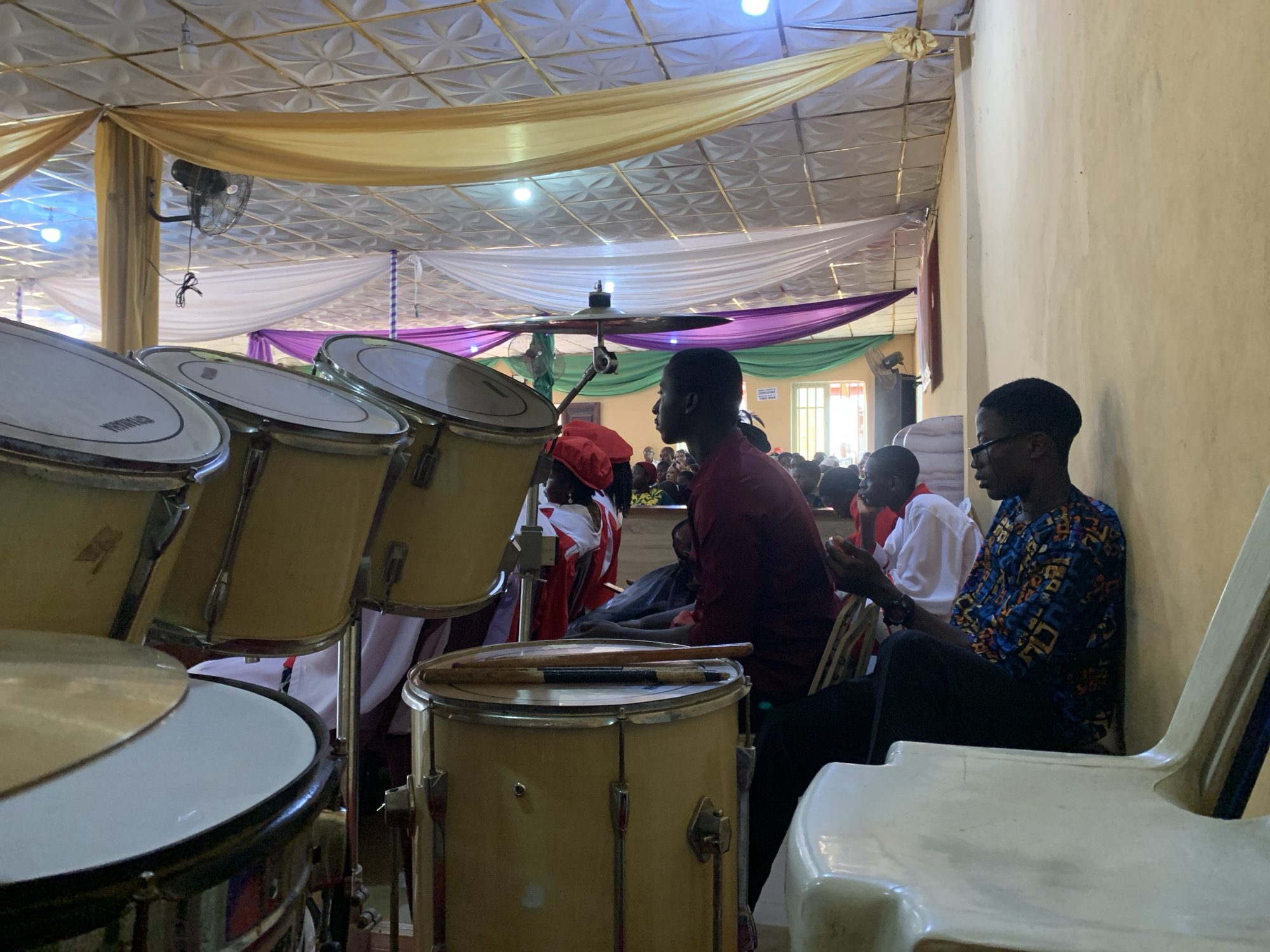 The image depicts a gathering of people inside a building with a decorated ceiling featuring draped fabrics of gold, white, purple, and green. Dominating the foreground is a yellow drum set. Behind the drums are seated individuals; one in a red shirt and another in a patterned blue shirt and glasses are clearly visible. Other people are partially visible in the background. The location is within a structure in Akpaka Forest Reserve, Nigeria.