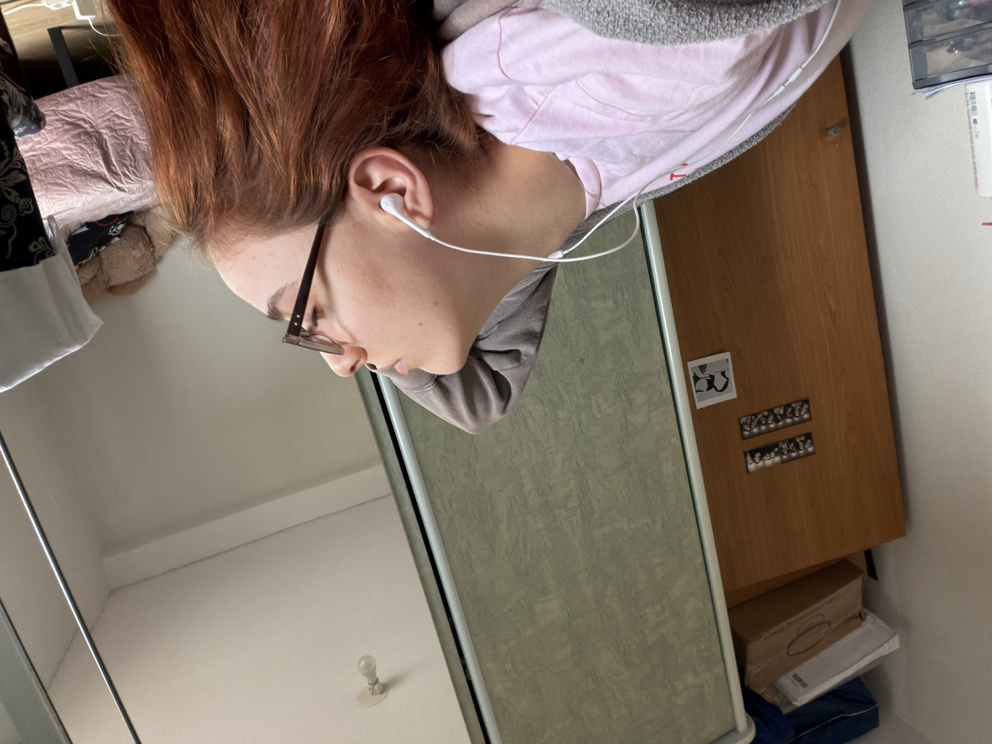 A candid, vertically oriented indoor shot captures a young individual, likely female, from the mid-torso up, seen in profile against a backdrop of their room. The person has reddish-brown hair and is wearing glasses, a light pink t-shirt under a grey hooded sweatshirt, and white wired earbuds in their right ear. Their head is tilted downwards, suggesting they are looking at something below the frame or focusing intently.
The setting appears to be a bedroom or dorm room. To the person's left, a bed with a pink textured duvet is partially visible, along with some dark patterned fabric and a white garment. To their right, a light brown wooden wardrobe or door dominates the background, featuring small black and white photo strips and a larger graphic or sticker on its surface. Beneath the wardrobe, several cardboard boxes are stacked, with one box showing "KIK" and "ANX" printed on it. A clear plastic organizer and white bottles or containers, one with a "BARCODE" label, are visible on a surface to the far right. The walls are white, and a bare light bulb is mounted on the ceiling, suggesting artificial illumination or a bright interior. The overall impression is a personal, lived-in space. The person's expression is neutral, consistent with being engrossed in listening or thought. No specific time of day or weather is discernible, and aside from the provided context, there are no explicit location cues for Strasbourg, France.