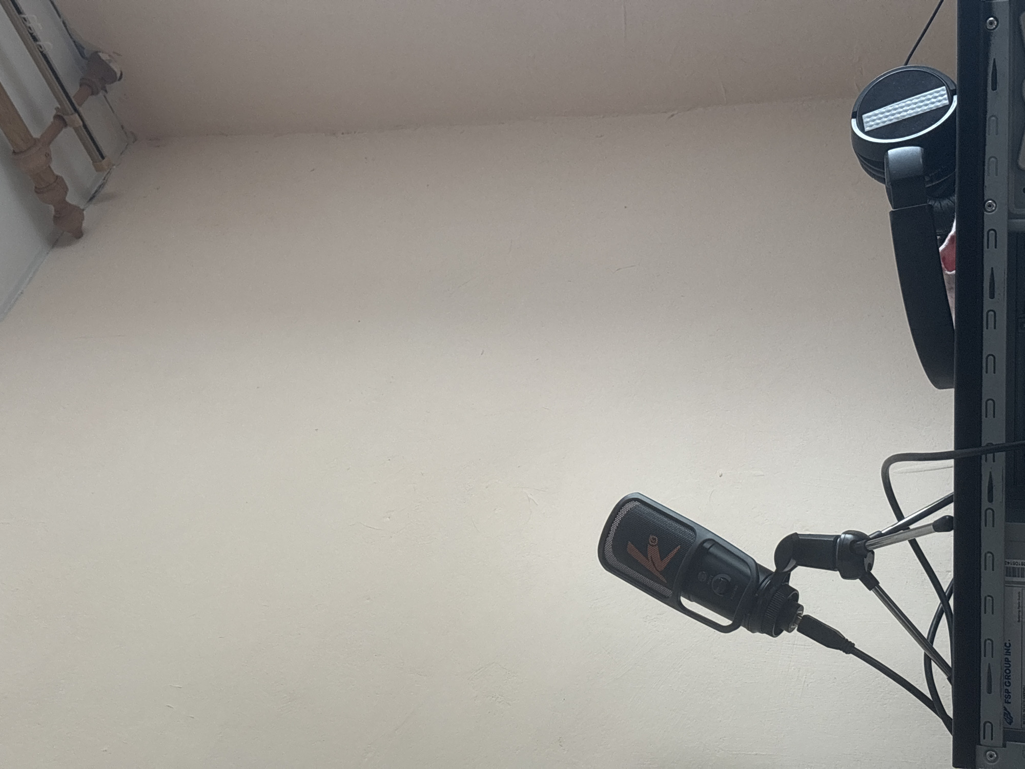 The image shows a close-up of a black microphone on a tripod, positioned on top of a computer tower. Next to the microphone, on the same surface, is a pair of black headphones. The background is a plain, light-colored wall, with some piping visible near the ceiling in the upper right corner. The scene appears to be indoors. There is no information in the image to indicate the location in the city of Kstovo, Russia.