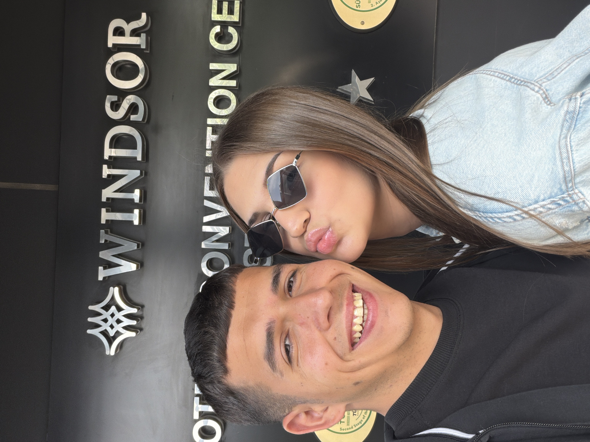 A man and a woman pose for a photo in front of the Windsor Convention Center in Bayrampaşa, Turkey. The man is in the foreground, smiling broadly, and wearing a black long-sleeved shirt. The woman is behind him, pouting, wearing sunglasses, and a denim jacket. The building sign behind them says "WINDSOR CONVENTION CENTER" vertically, with a round logo to the right.