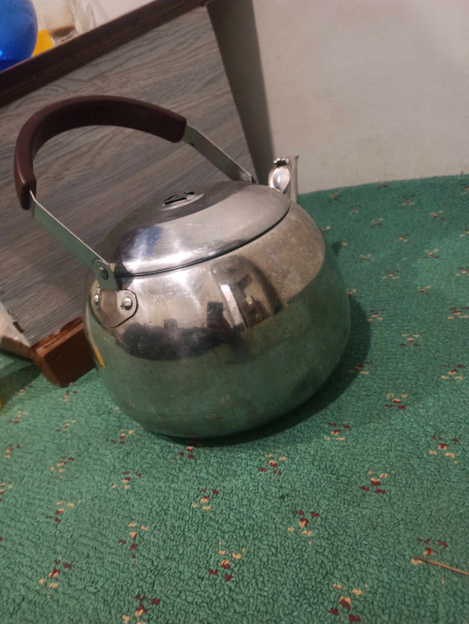 The image shows a close-up, angled view of a stainless steel tea kettle with a brown handle, resting on a green, textured surface with a subtle pattern of small, scattered red and off-white dots. The kettle is positioned to the left of the frame, taking up a significant portion of the foreground. Its surface is reflective, showing distorted reflections of the surrounding environment, including what appears to be a portion of a wall and possibly a window or light source. To the left of the kettle, a wooden surface is visible, with a blue object and a clear glass or plastic container partially visible in the upper left corner. The background consists of a plain, light-colored wall. The scene appears to be indoors, possibly in a domestic setting. There are no people visible in the image. The location of this scene in Sumedang, Indonesia, cannot be determined from the visual information provided in the image.