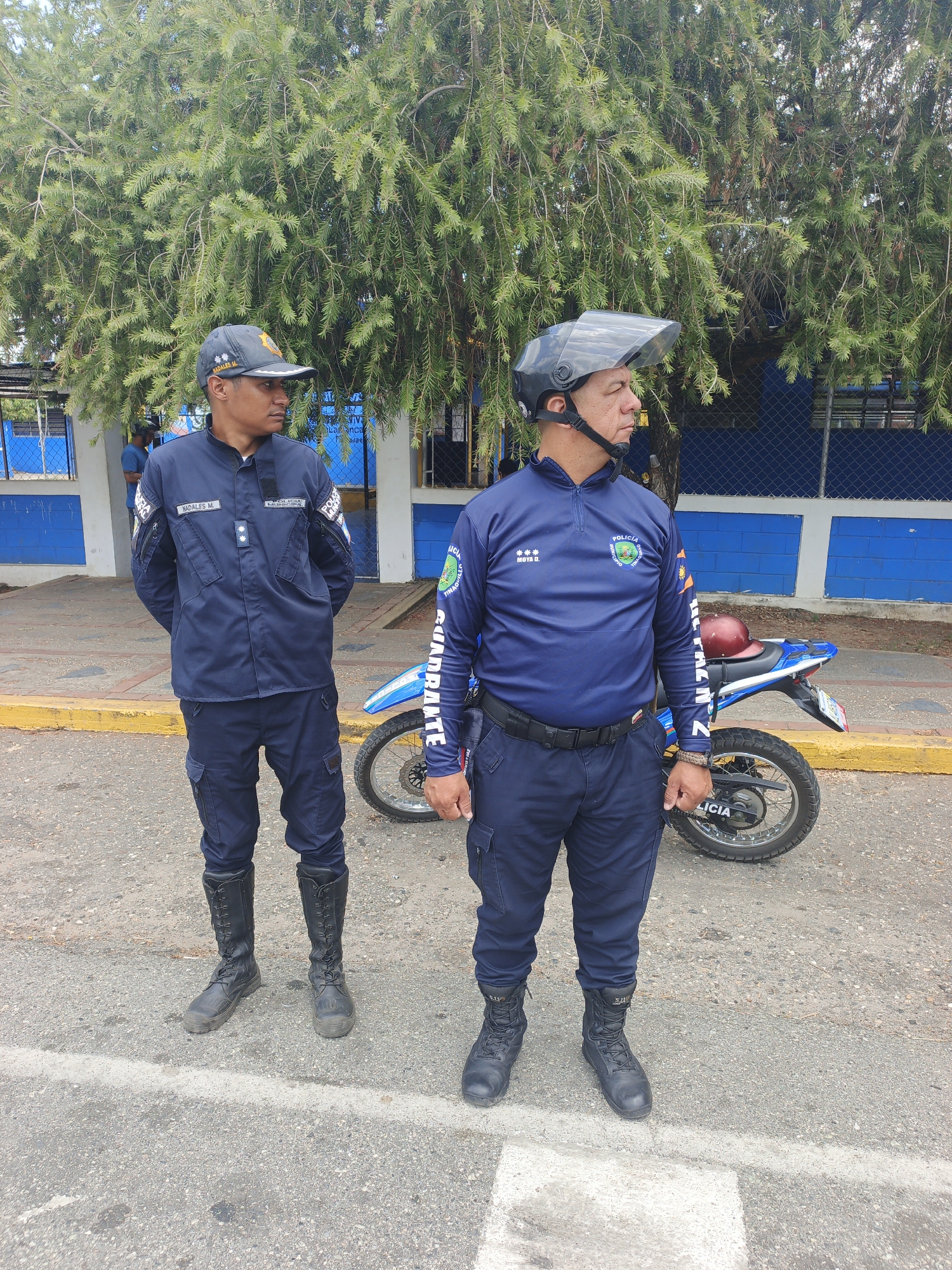 The image shows two uniformed police officers standing on a street in Tinaquillo, Venezuela. A motorcycle is parked behind them, next to a building. The officer on the left is wearing a cap, jacket, and tall boots, with arms crossed. The officer on the right is wearing a helmet with a clear visor, a long-sleeved shirt, pants, and shorter boots. They are both looking off to the side. A tree is visible behind the officers.