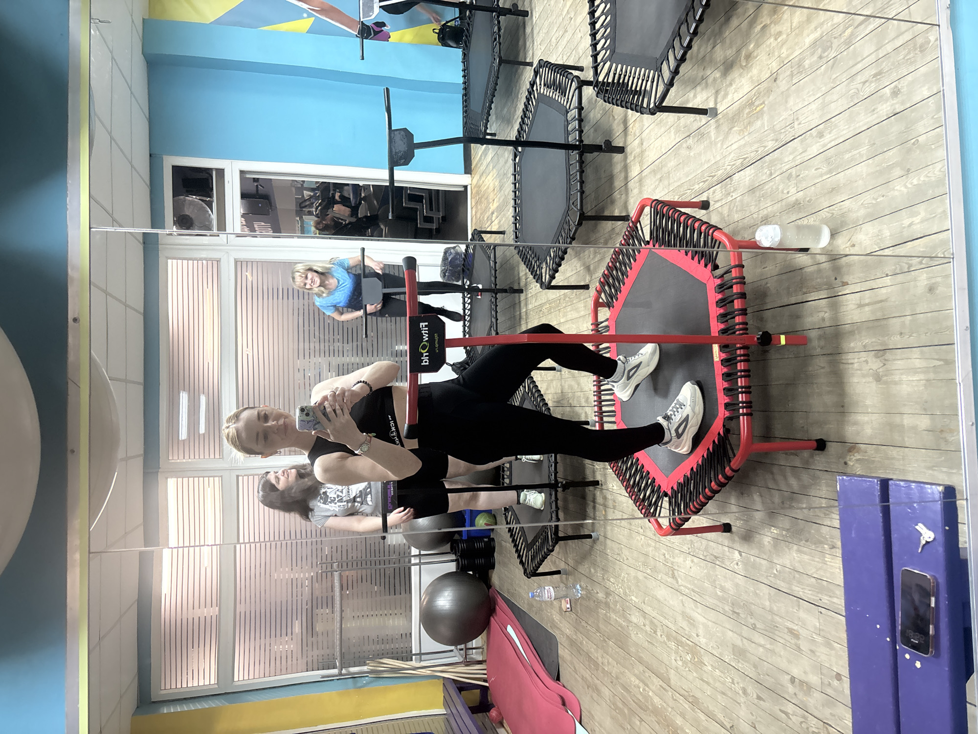 A mirrored view of a woman in a gym setting is captured. She is dressed in black athletic wear and white sneakers, standing on a small trampoline with a red frame. The woman is holding a phone and appears to be taking a selfie or video. Her reflection is visible in the mirror behind her. A woman in a blue shirt appears to be nearby. In the background, there are multiple similar trampolines and other gym equipment. The gym has a wooden floor and blue, yellow and pink walls. A water bottle is on the floor next to the trampoline. A smart phone is attached to a blue cabinet in the lower right corner of the picture. Based on the background, it is likely that this gym is in Saint Petersburg, Russia.