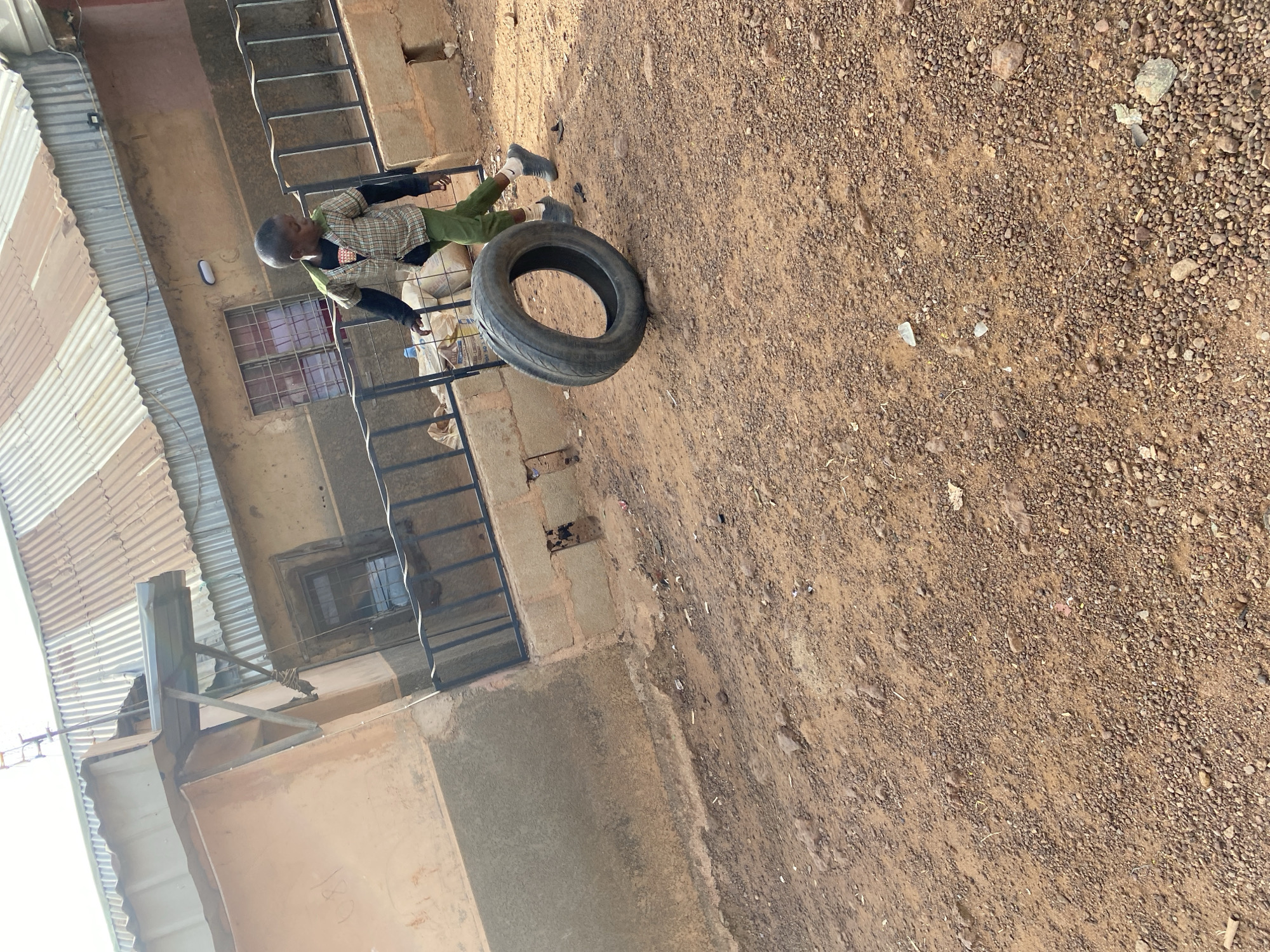 The image depicts an outdoor scene in Bukuru, Nigeria, featuring a young boy and a building with unpaved ground. The main subject is a young boy, appearing to be of school age, dressed in a plaid long-sleeved shirt, green trousers, dark shoes, and white socks. He has closely-cropped dark hair or is wearing a dark cap. He stands upright, facing towards the left side of the frame, with his hands near a black metal railing.
A used black vehicle tire lies on its side on the ground close to the boy. The ground is unpaved, composed of reddish-brown dirt and small gravel, indicating a dry, outdoor environment. A cropped view of the ground reveals small pieces of broken glass.
In the background, there is a multi-story building with light brown or tan walls. It features a window secured with metal grates and a simple light fixture on the wall. A prominent element on the left is corrugated metal roofing, likely belonging to an adjacent structure. Behind the metal railing where the boy stands, several white sacks or bags are visible, possibly containing supplies. Faint, handwritten numbers "180" can be seen on a section of the building wall.
The scene is illuminated by natural daylight, suggesting a clear day, possibly late morning or early afternoon. The boy's expression is neutral, and he appears to be standing or observing rather than actively engaged in a specific event or play at this moment. The setting reflects a functional, everyday environment typical of semi-urban or rural areas in the region.