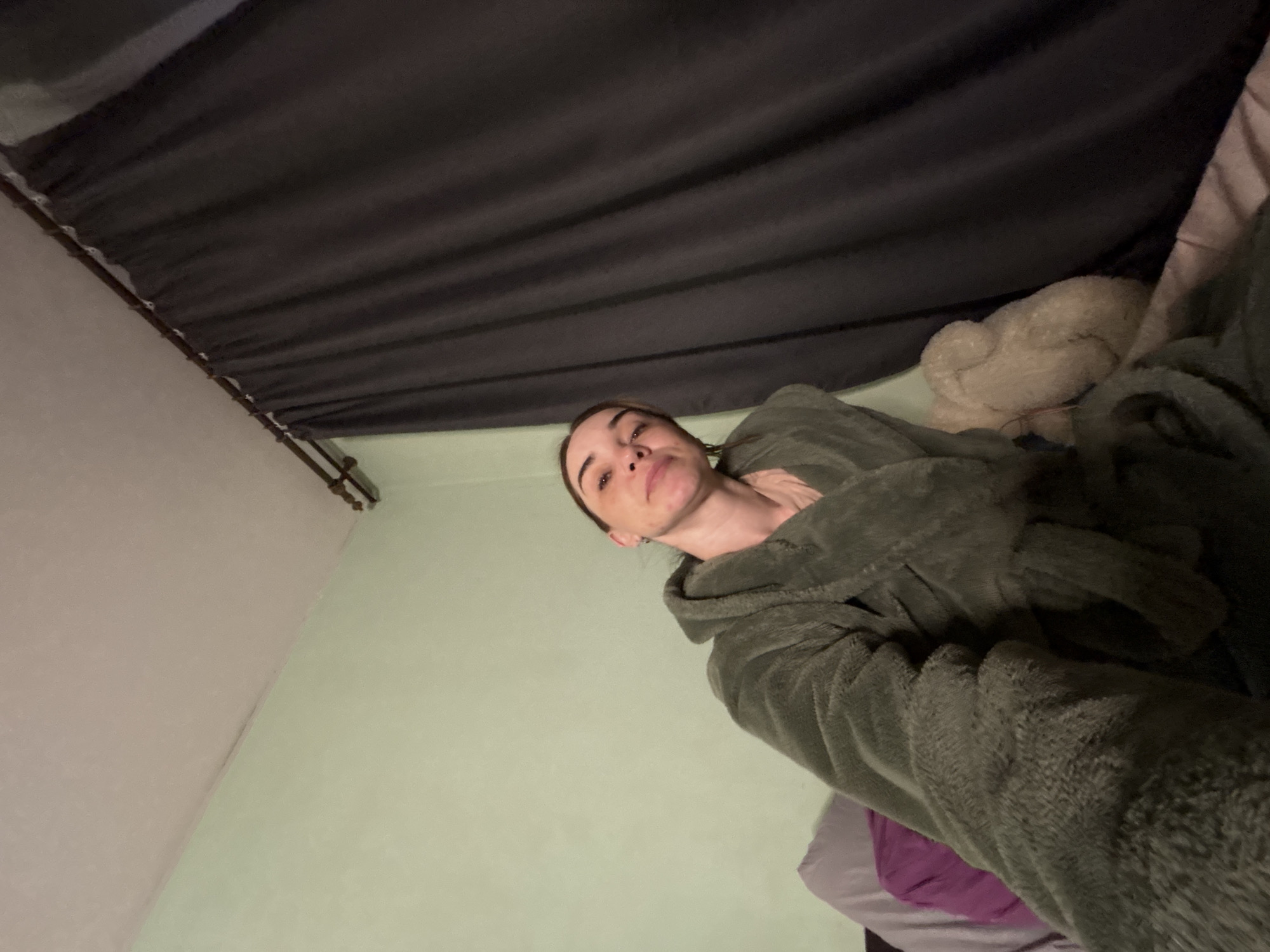 A woman wearing a hooded robe is lying in bed. A gray curtain hangs from the ceiling. The walls are light green and white. A stuffed animal and purple bedding are also visible. The location is Unknown, Unknown.