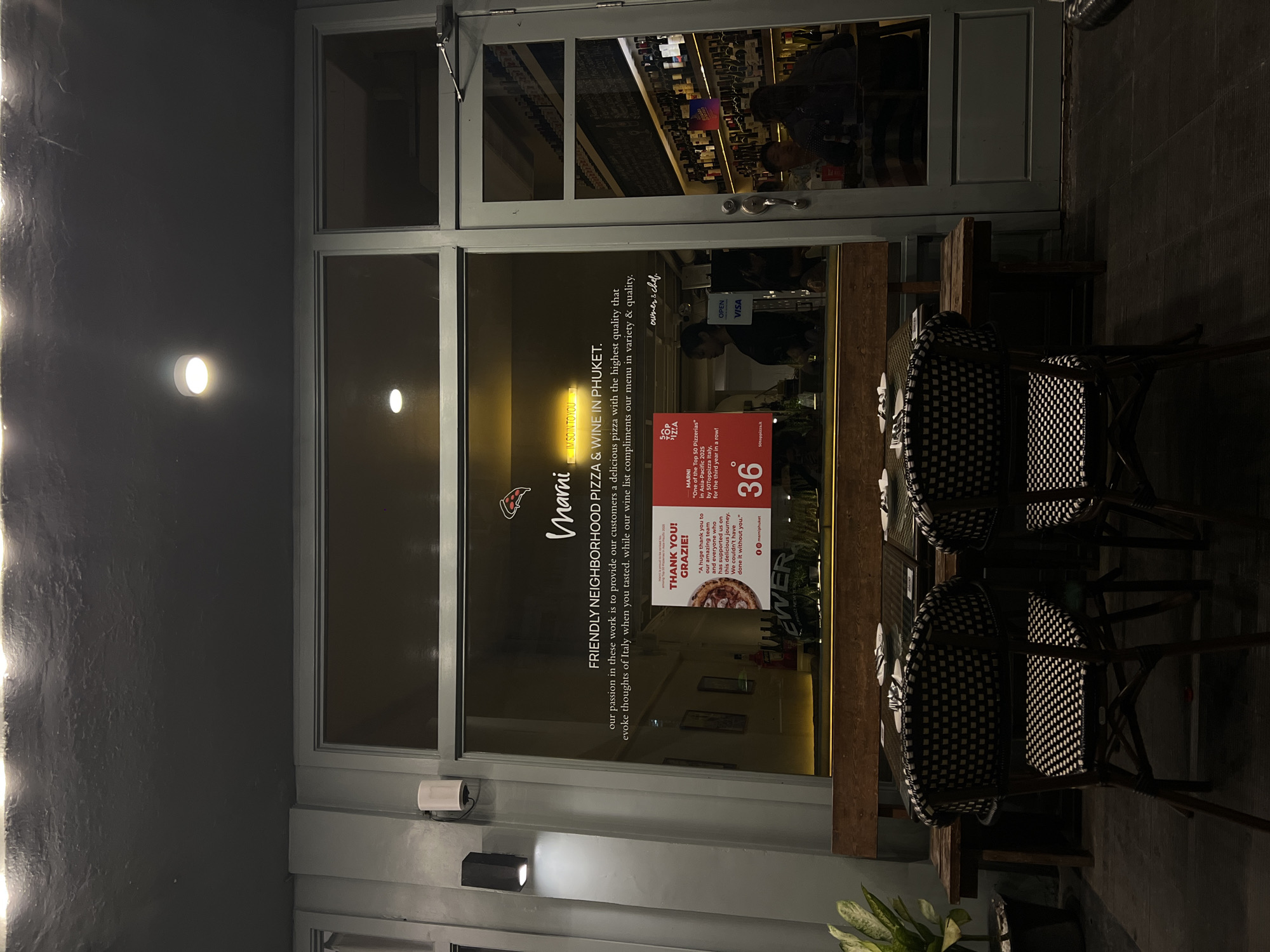 The image depicts the storefront of "Marni," a friendly neighborhood pizza and wine restaurant located in Phuket. The signage on the glass states that the restaurant serves delicious pizza with the highest quality ingredients and aims to evoke thoughts of Italy, and they were included in the Top 50 Pizzerias in Asia-Pacific 2023 by 50Toppizza Italy. A thank you sign acknowledges the support of the team and customers. Two chairs and a table, set up for outdoor dining, are visible outside the entrance.