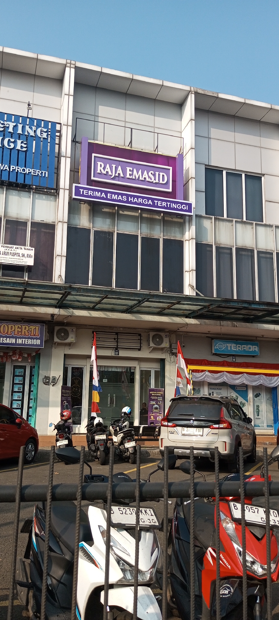 This is a daytime, eye-level, medium shot of the exterior of a commercial building with shops on the ground floor. The building is modern with white cladding and large windows. Several shops are visible, including one with a purple sign that reads "RAJA EMAS.ID" and another with a blue sign that reads "ISTINGE". There are also signs for "TERADA" and "PROPERTI". In front of the shops, several motorcycles are parked. A silver SUV is parked in front of the "TERADA" shop. The sky is clear and blue. The location is Lambangjaya, Indonesia, as indicated by the signs. There are no people clearly visible in the image. The scene appears to be a commercial street in a suburban or urban area of Lambangjaya, Indonesia.