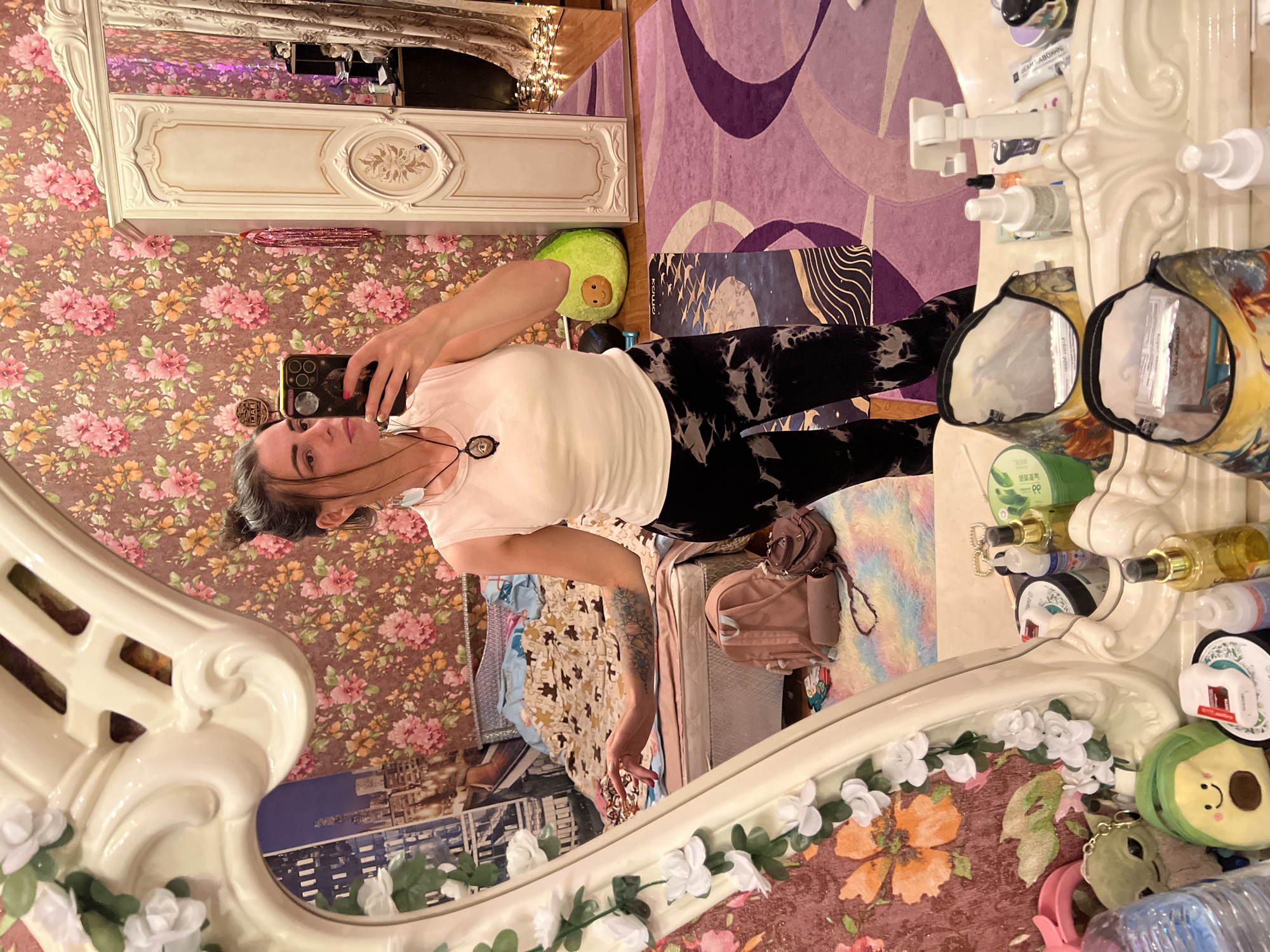 A young woman stands in front of a mirror, taking a selfie. She has dark hair pulled up in a bun and is wearing a white tank top and black and white tie-dye leggings. She is wearing a necklace with a dark pendant. In the mirror, her reflection shows her in a room with floral wallpaper and a bed in the background. There are several decorative objects visible in the reflection, including a large green avocado-shaped plush toy and a pink backpack. The room appears to be a bedroom. The woman is positioned in the center of the frame, with the mirror taking up the majority of the image. The background is out of focus but suggests a busy and somewhat cluttered environment. The overall impression is of a candid personal moment captured in a domestic setting.
