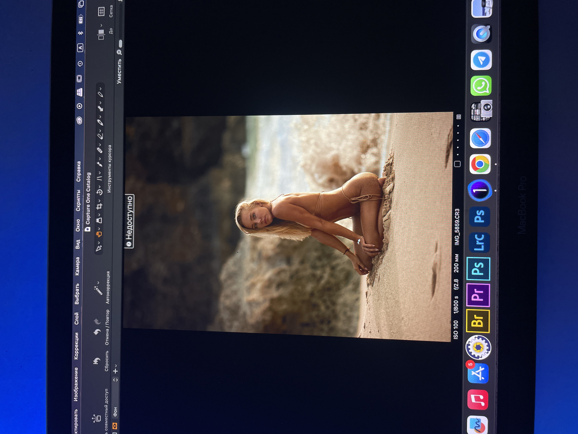 A photograph of a MacBook Pro screen shows the Capture One photo editing software open with an image of a woman posing on a beach. The woman is blonde, wearing a small bikini, and kneeling in the sand. The image is well-lit, and the background shows ocean water and rocks. The MacBook's dock is visible at the bottom of the screen, showing a variety of applications. The menu bar at the top shows options in Russian. Kazan, Russia is not present in the image.