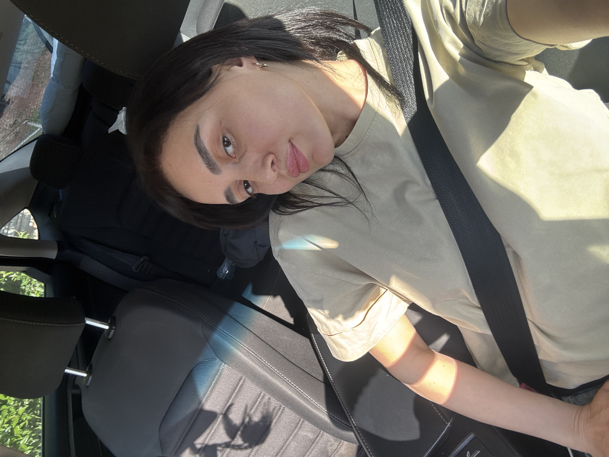 A woman with dark hair is inside a car, wearing a tan t-shirt and a seatbelt. The car appears to be parked; it is likely in Tbilisi, Georgia, because the window is visible and the background includes the typical trees and vegetation found in residential areas of the city. The car's interior consists of dark grey seats. The woman's face is partially lit by sunlight.