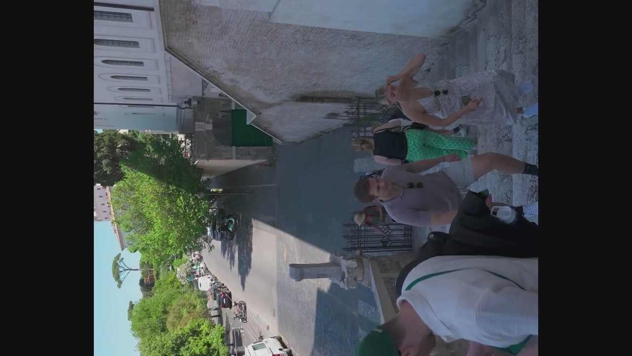 The image shows a group of people walking up a stone staircase, suggesting a public area or a historic site, possibly in a European city given the architecture and the presence of scooters and cars on the street below. The staircase is bordered by a brick wall on the right and a metal gate on the left. The people are dressed in casual summer clothing. In the background, a street with parked vehicles and trees is visible, with buildings and a distinct tree that appears to be a pine tree in the distance. The lighting suggests it is daytime. The presence of a white van and multiple cars indicates an urban or semi-urban environment. The overall impression is of a tourist or pedestrian area. The question states the location is Morlupo, Italy, which is a commune in the Metropolitan City of Rome Capital. The scenery is consistent with a location in or near Rome, which is known for its historic architecture and cypress trees.