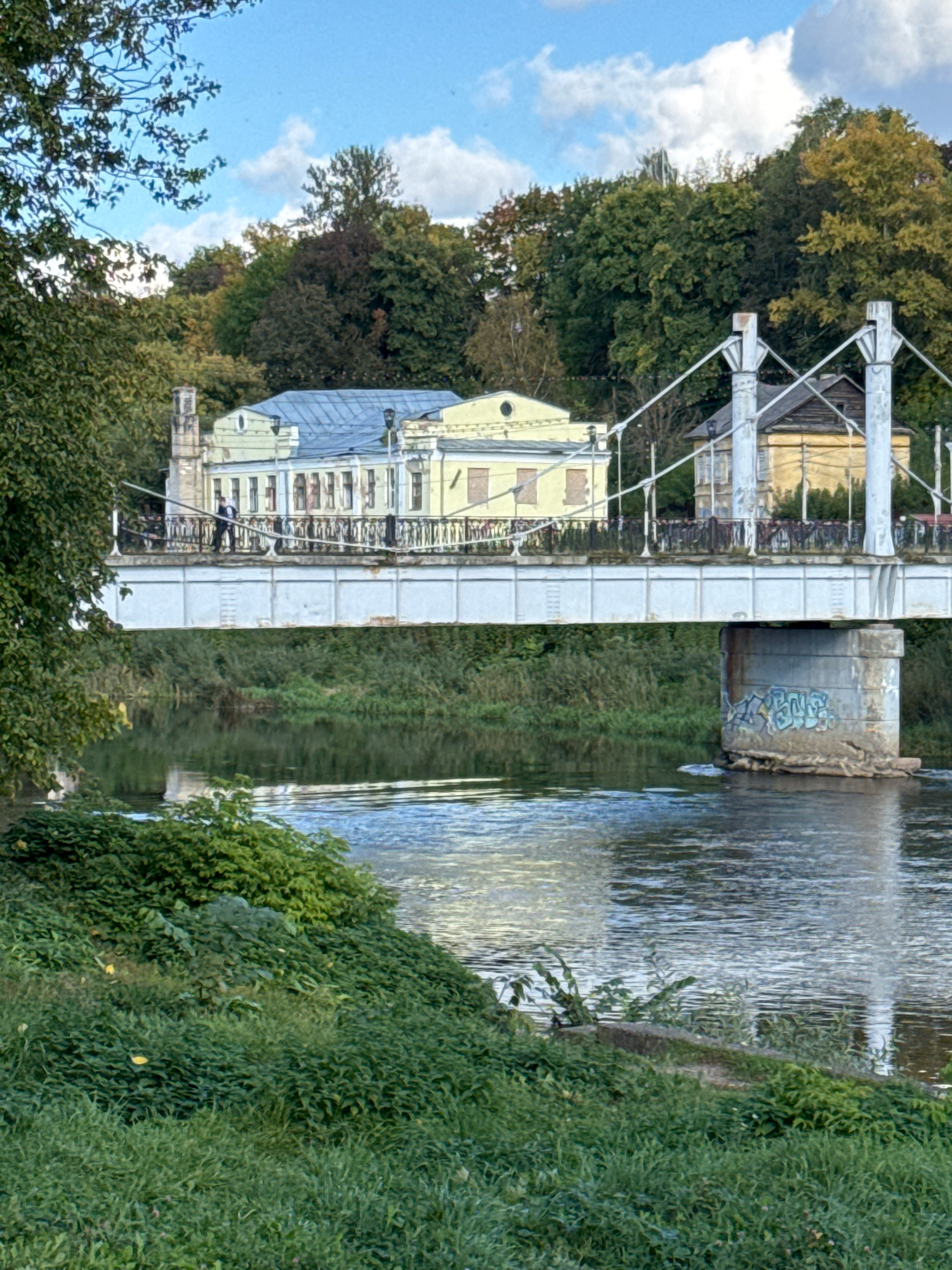 A sunny day scene in Torzhok, Russia, features a bridge spanning a wide, calm river. The bridge has a metal railing and a pedestrian walkway, with two white suspension towers visible. A person dressed in a dark suit and tie stands on the walkway, facing away from the camera. On the far bank of the river, behind the bridge, are several buildings with yellow and light blue facades, surrounded by lush green trees and foliage. The river's surface is calm, reflecting the sky and surrounding greenery. The bridge's concrete support in the foreground is adorned with graffiti. The scene is set on a bright, clear day with scattered white clouds in the blue sky.
