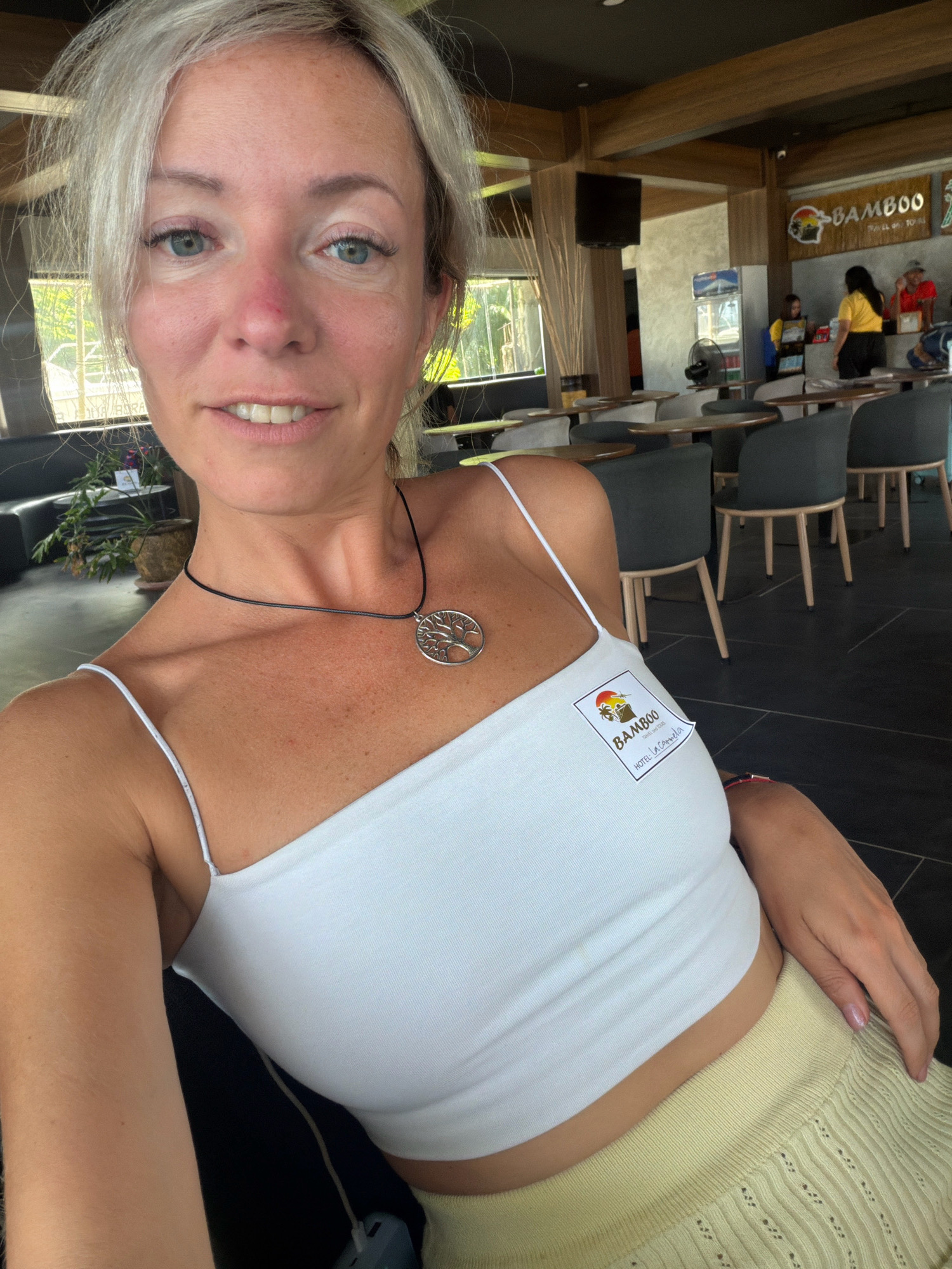 A fair-skinned woman with blonde hair and blue eyes is captured in a selfie taken indoors. She is wearing a white spaghetti strap top and a light yellow knitted skirt. A silver tree of life pendant hangs from a black cord around her neck. A sticker with a logo featuring palm trees, the text "BAMBOO", and "HOTEL LA CARMELA" is affixed to the left side of her top. The background reveals a restaurant or cafe setting with wooden beams, tables, and chairs. In the distance, other patrons and staff are visible. The lighting suggests it is daytime. The sticker indicates the location is Hotel La Carmela in Malay, Philippines.