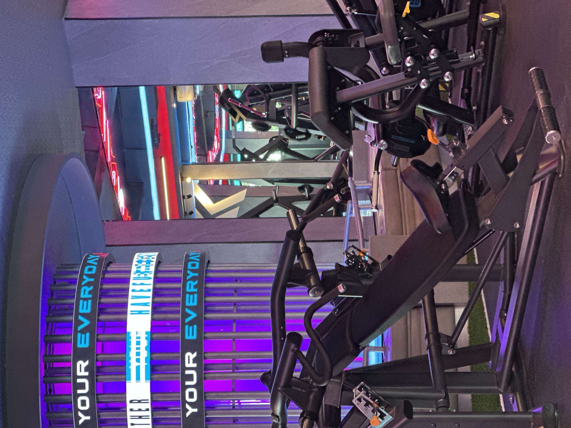 The image captures a scene within a brightly lit fitness center in North Kuta, Indonesia. Several pieces of black weight training equipment dominate the foreground, arranged in a linear fashion. These machines have a complex arrangement of bars and levers, suggesting different exercise functionalities.
In the background, there is a large cylindrical structure with neon signs wrapping around it. The signs read "YOUR EVERYDAY", with the text vertically aligned, indicating the fitness center's motto or brand statement. The color scheme of the signs is predominantly blue and white against a black background, illuminated with blue and purple lights shining through the bars surrounding it.
A glimpse of another area with neon signs and a grey floor can be seen further back, offering a peek into other areas of the gym. The overall environment is modern, and the lighting contributes to a high-energy atmosphere typical of fitness centers.