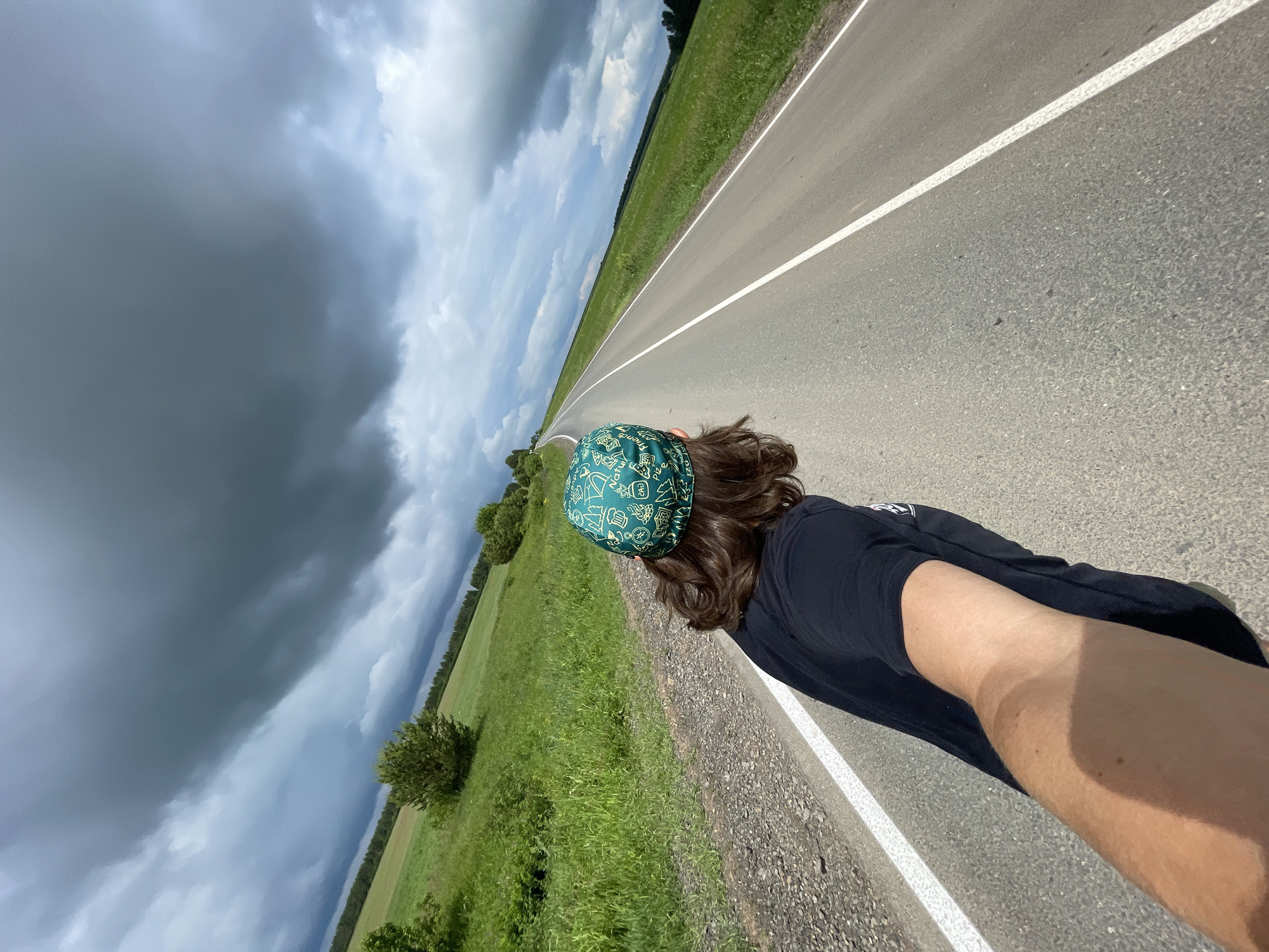 A diagonal, high-angle shot captures a person from behind, facing a long, straight asphalt road extending into the distance. The road features a clear white line marking its edge. On both sides, expansive green fields stretch towards a horizon defined by distant lines of trees.
The subject has medium-length brown hair and wears a dark green bandana patterned with numerous light-colored cartoon-like icons, including pizza slices, ghosts, skulls, mushrooms, flames, and the word "FRIENDS" repeated. They are dressed in a black short-sleeved t-shirt. Their right arm is visible, showing a noticeable tan line, suggesting exposure to sunlight.
The sky overhead is dramatic and dynamic. The left side of the frame is filled with heavy, dark storm clouds, indicative of an approaching or recently passed rain event. In contrast, the right side of the sky shows lighter, though still overcast, clouds. The overall scene suggests an open, rural landscape under changeable weather conditions, likely during the daytime. The person appears to be traveling or standing by the road, observing the vast environment.