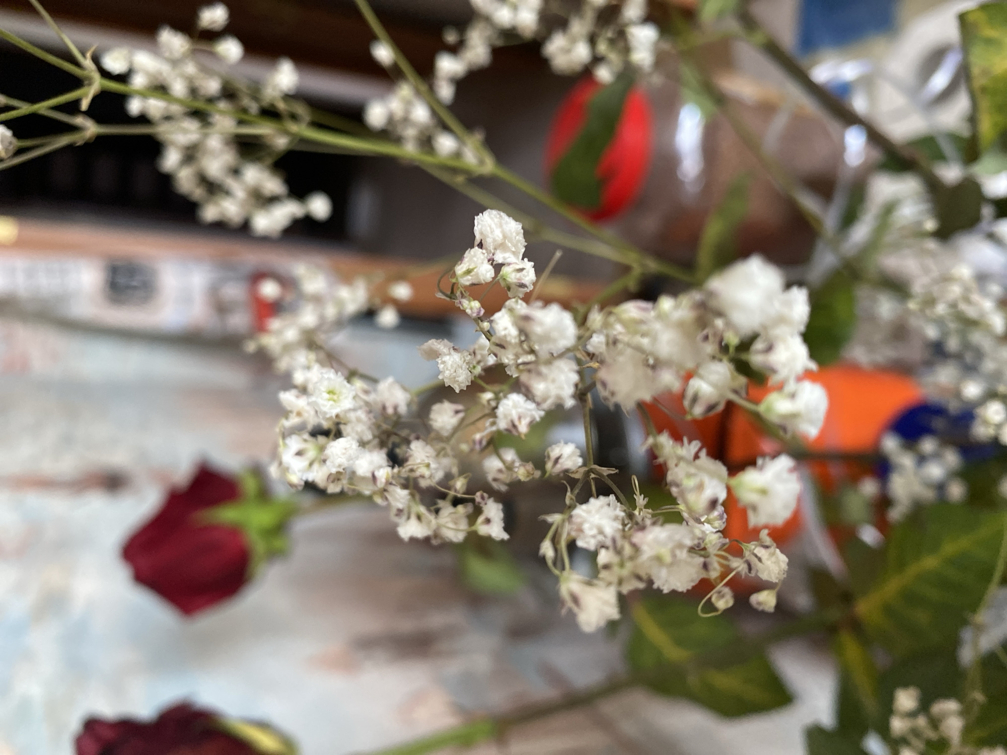 The image is a close-up, slightly out-of-focus photograph of a floral arrangement. The primary subjects are small white flowers, possibly baby's breath, which are clustered on delicate green stems. In the background, out of focus, are two dark red rose buds. The environment appears to be indoors, with a textured, possibly brick or stucco wall visible behind the flowers. To the right, a glass jar with a red lid and some brown contents is partially visible. The overall impression is of a still life with a focus on delicate textures and muted colors. The image does not provide any information that would allow for its location to be determined to the city of Molodizhne, Ukraine.