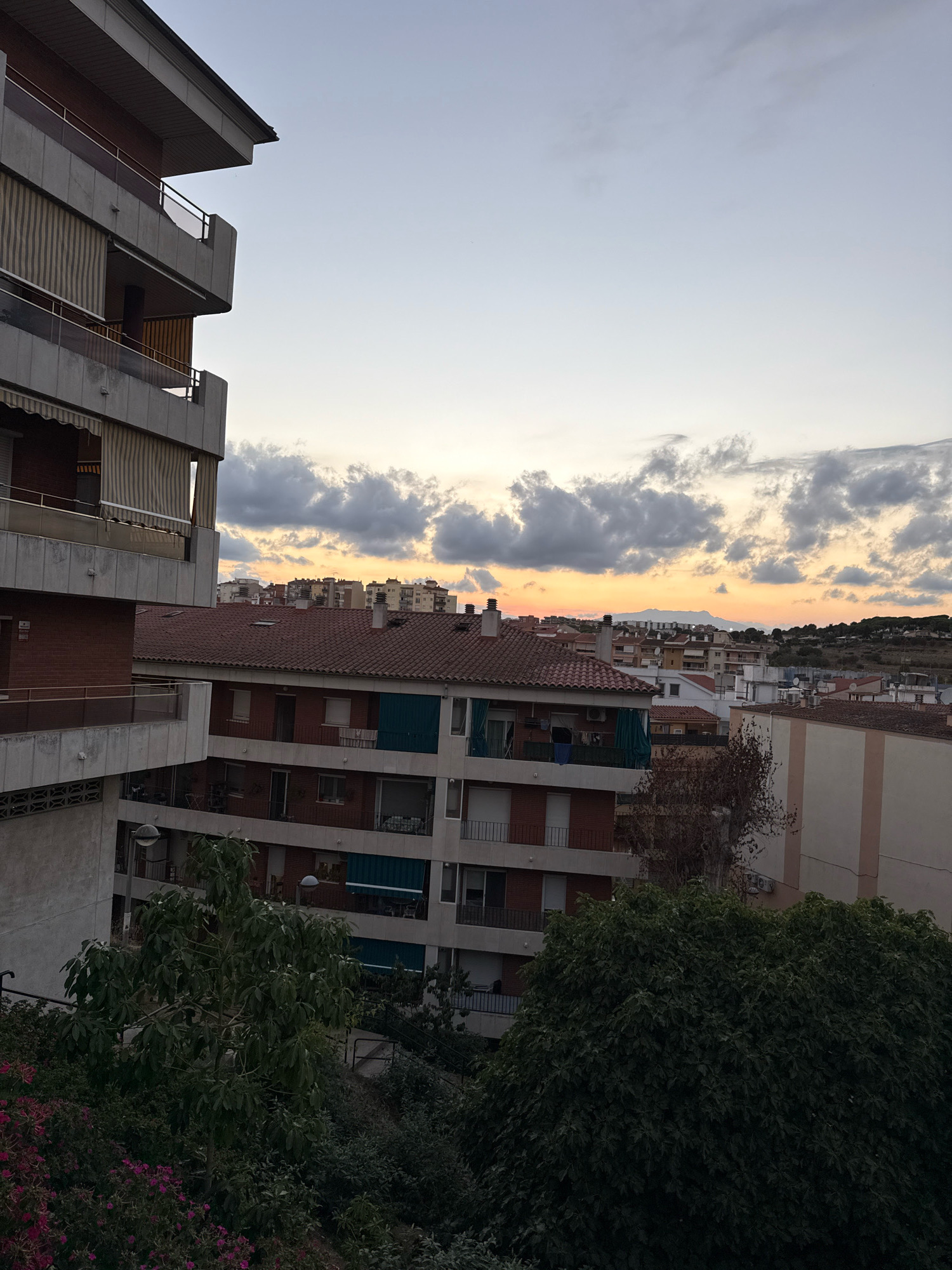 The image captures an urban landscape in Blanes, Spain, at dusk. The foreground is dominated by the balconies of a residential building, characterized by concrete structures, glass railings, and beige striped awnings. Below, lush green trees and vibrant pink flowers create a dense natural buffer.
In the middle ground, a series of multi-story apartment buildings with red tiled roofs and visible windows are arranged closely together. Some buildings feature balconies with greenery or clothing drying. The buildings extend towards the background, forming a cityscape.
The sky above is a gradient of soft blues and grays, transitioning to a warm orange and yellow hue near the horizon, indicative of sunset. Scattered, dramatic clouds dot the sky, adding texture to the scene. In the far distance, a faint silhouette of hills or mountains is visible against the twilight sky. The overall impression is one of a residential area in a coastal town as day transitions into night.