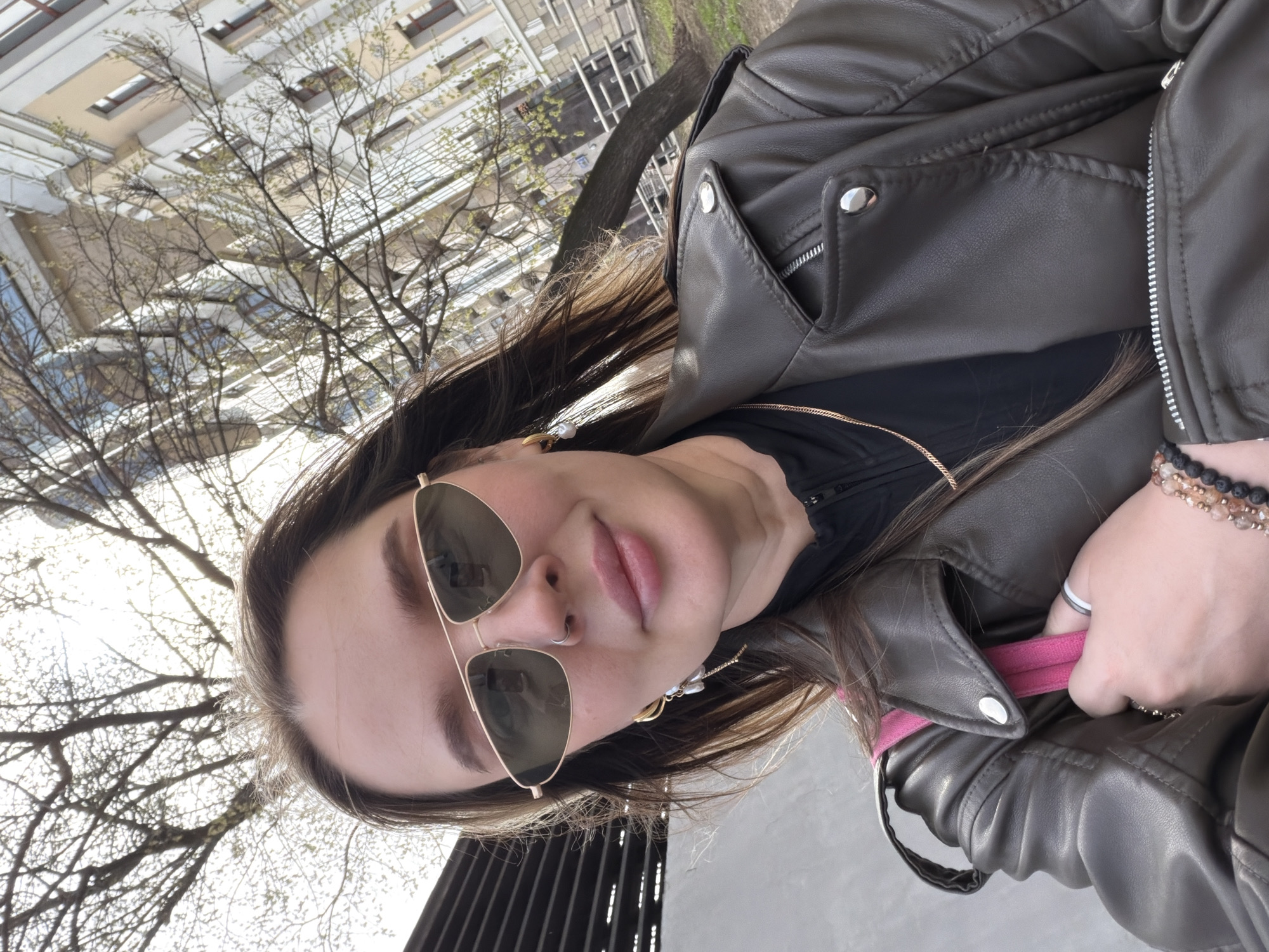 A young woman wearing aviator sunglasses, a brown leather jacket, and beaded bracelets takes a selfie in Moscow, Russia. Bare tree branches and a modern building can be seen in the background. She is also wearing a black top with gold zipper detailing and delicate earrings. Her face is partially obscured by the sunglasses. Her hair is long and brown, with blonde highlights. A pink strap is visible.