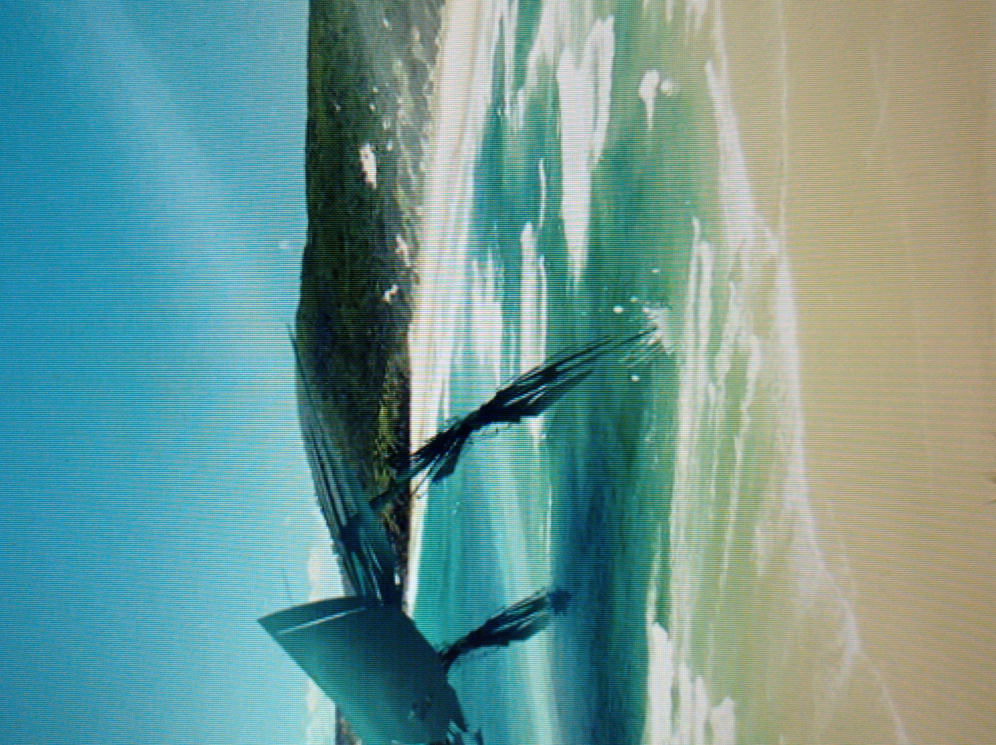 The image depicts a futuristic or alien spacecraft with a sleek, metallic design, partially submerged in turquoise water near a sandy beach. The spacecraft has multiple pointed appendages extending into the water, creating ripples and splashes. In the background, a lush green hill slopes down towards the coastline, under a clear blue sky. The scene appears to be a digitally rendered or composited image, as it lacks the naturalistic detail of a real photograph. There are no people visible in the image, and no identifiable landmarks or context to suggest a specific location in Mukachevo, Ukraine. The image's resolution is low, with visible pixelation, suggesting it may be a low-quality screenshot or a simulated environment.