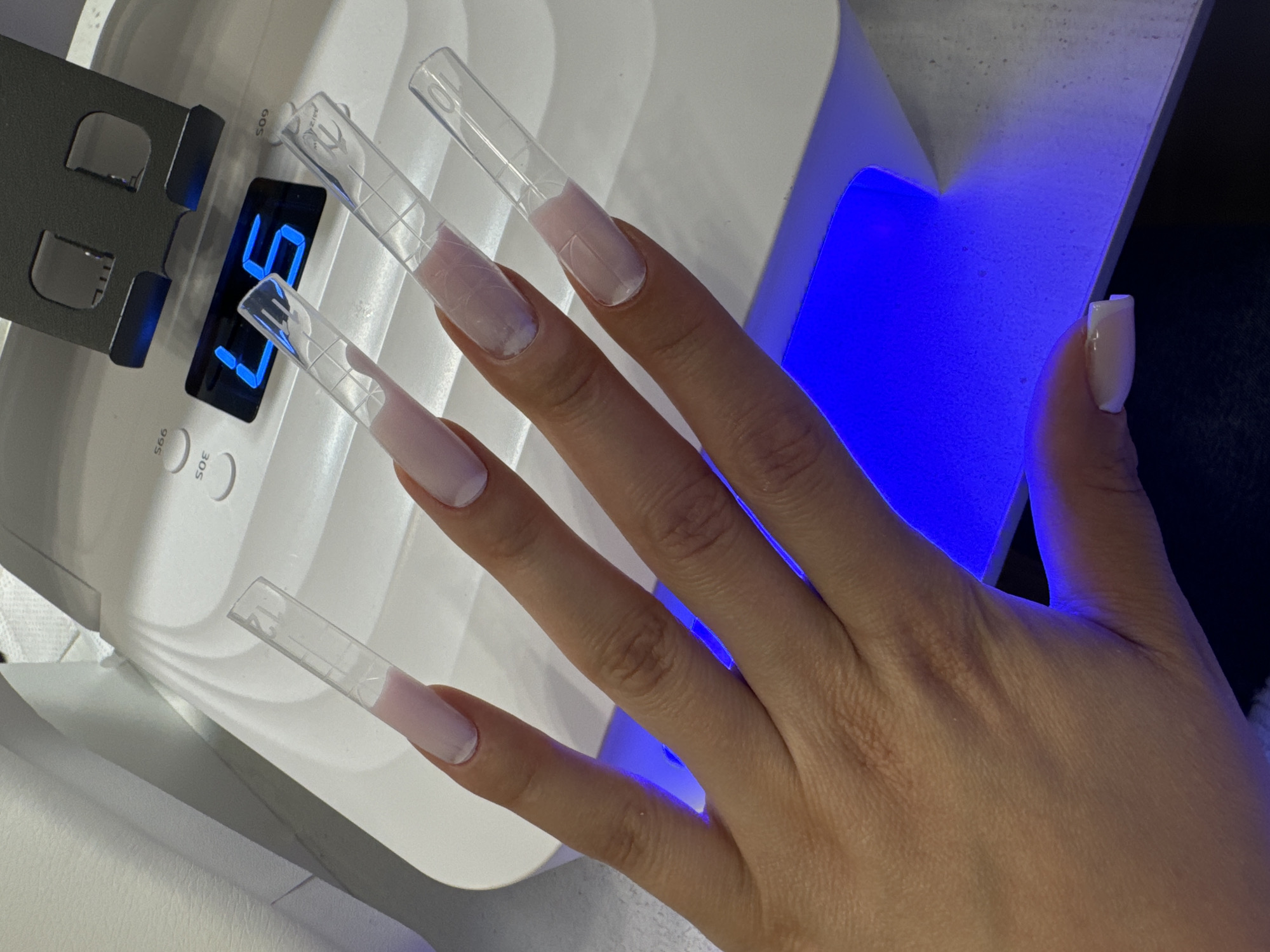 A person's hand is positioned over a white nail curing lamp. The hand has five fingers, and four of the nails have clear plastic forms attached to them, extending the nail length. The nails are painted with a light pink color. The lamp's digital display shows the number "57." The environment appears to be indoors, with a dark background visible to the right of the lamp. The location is not indicated within the scene, so there is no way to tell whether it is in Vniissok, Russia.