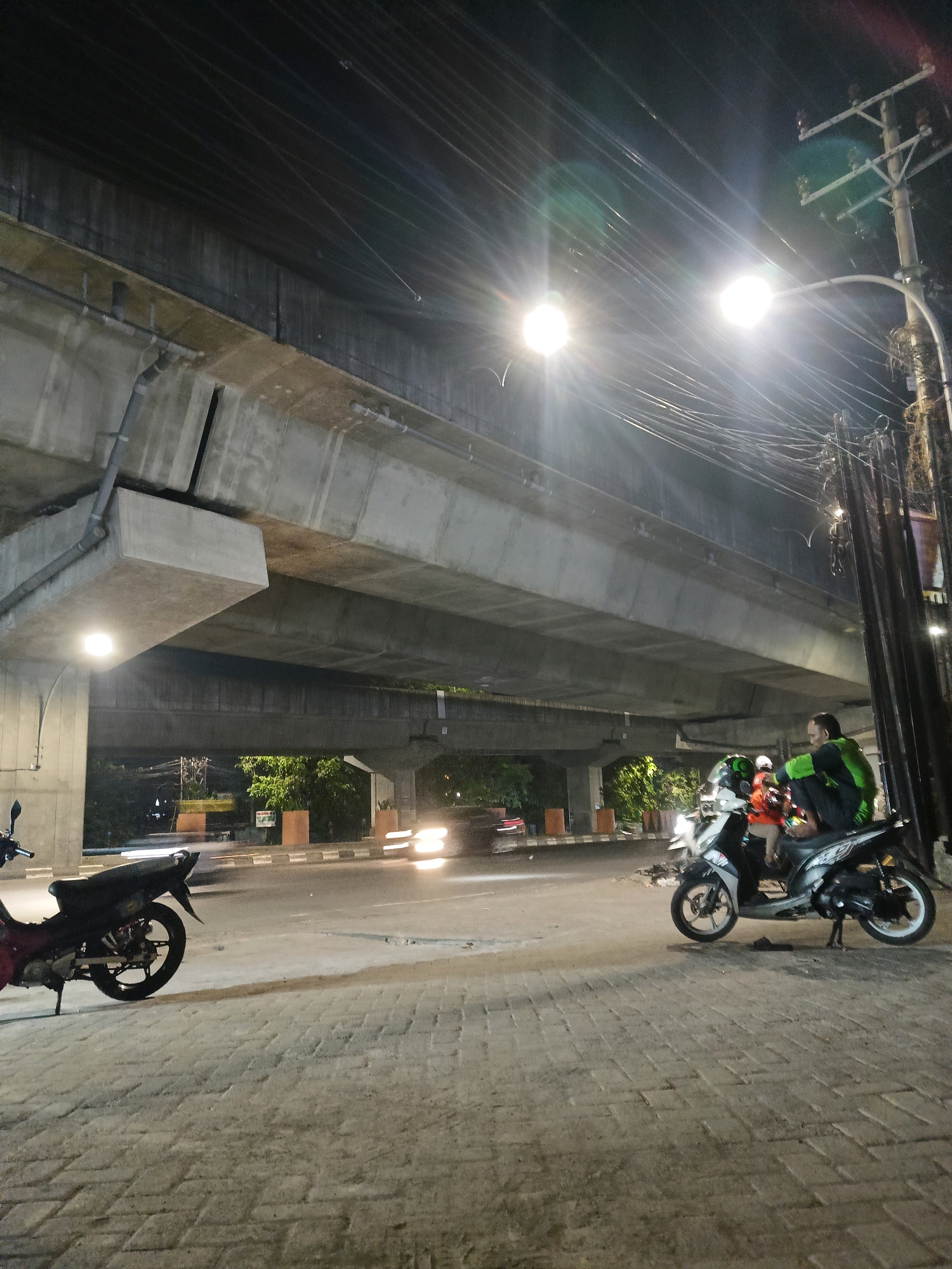 This is a nighttime photograph taken from street level, looking up at an elevated concrete overpass. The scene appears to be in Makassar, Indonesia. Two motorcycles are parked on a brick-patterned sidewalk in the foreground. On the right, a person wearing a green and black jacket is seated on a motorcycle, facing away from the camera and looking towards the right. Another motorcycle is parked on the left side of the image, partially visible. The road beneath the overpass is illuminated by streetlights and vehicle headlights, with some motion blur indicating traffic. Dense overhead utility lines are visible against the dark sky, crisscrossing above the overpass and streetlights. Lush green trees are visible behind the overpass in the background, dimly lit.
