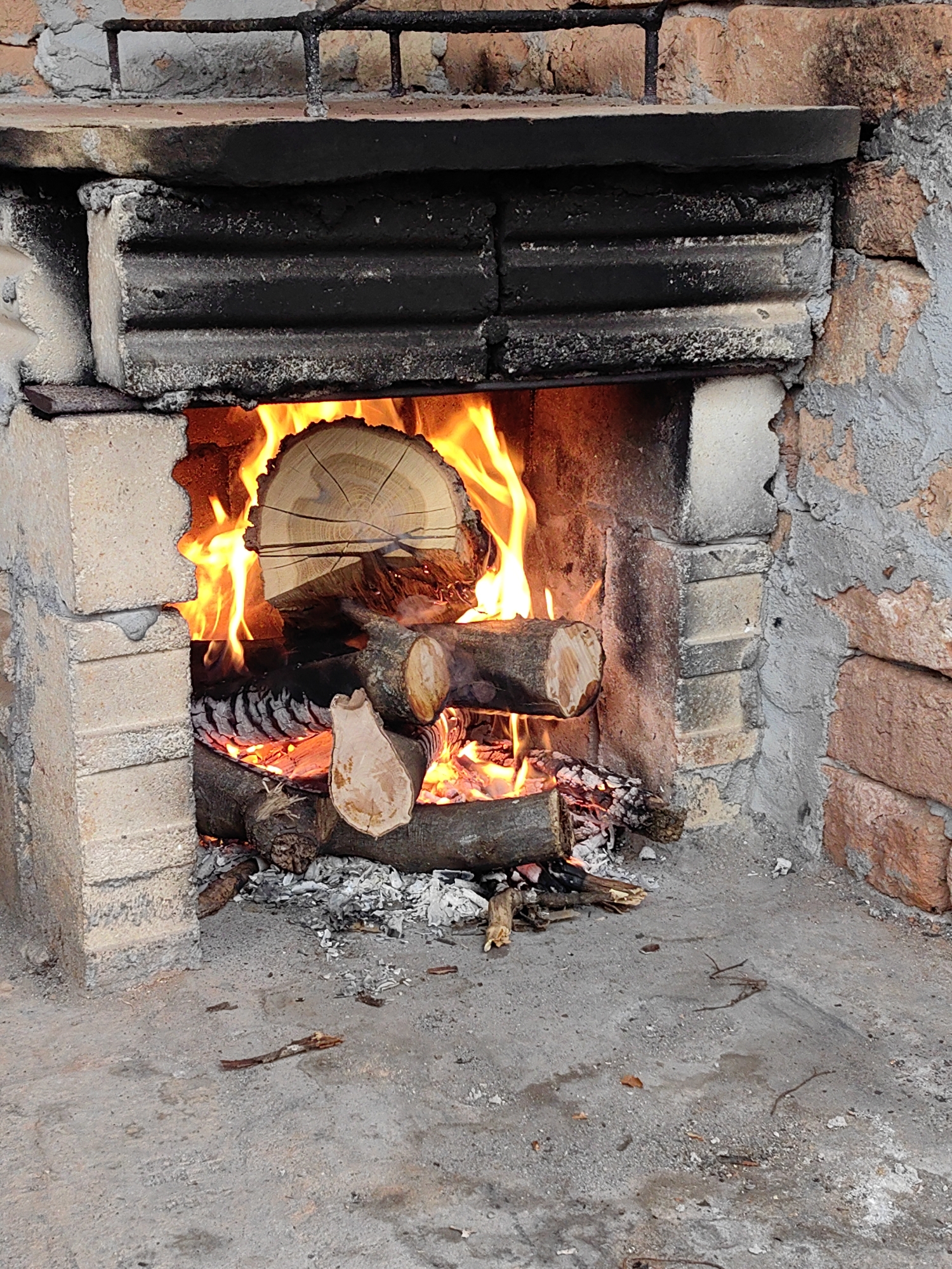 The image shows a brick fireplace in Dobrodan, Bulgaria, with a fire burning inside. Logs are arranged within the firebox, flames rising around them. Ash and burnt remnants lie below the logs. The surrounding brickwork shows signs of wear and soot. The ground in front of the fireplace is concrete.