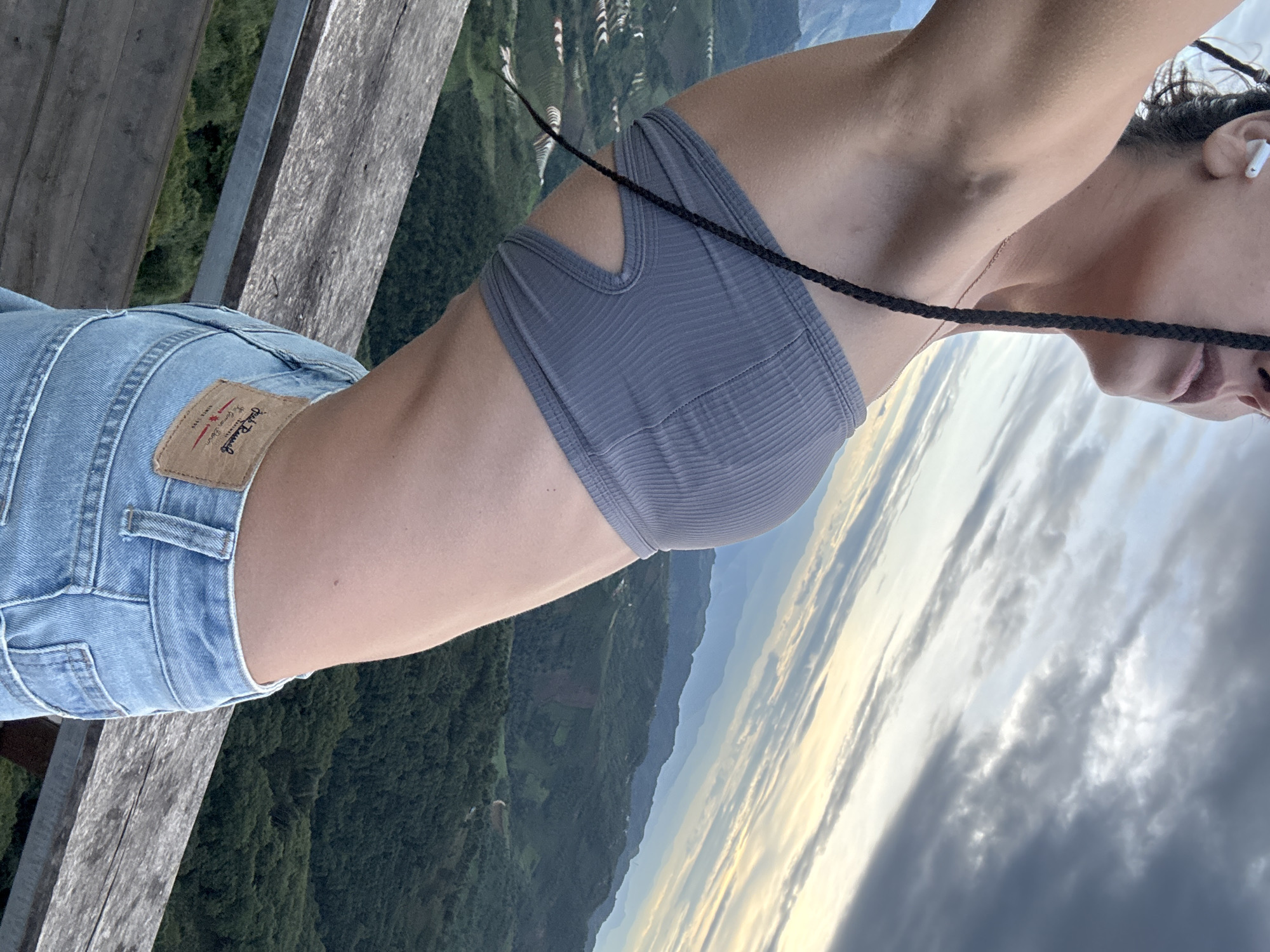 An individual is depicted from the upper torso to the hips, viewed from a slightly low and left-angle perspective. The subject's right profile is visible, looking upwards and to the right, with a white earbud in their right ear and a dark braided hair extension or braid draped over their right shoulder. They are wearing a grey, ribbed, strapless crop top featuring a cut-out design on the right side, exposing the ribcage. A thin gold-colored necklace is visible. The lower portion of the subject's torso is exposed, revealing skin with a small mole on the right side of the abdomen. Light blue denim jeans with a visible brown leather patch on the back right pocket area are worn; the patch features text including "Jack Russell," "Premium Quality Denim," and "SINCE 1999." The individual stands before a rustic wooden railing or ledge. The background reveals an expansive mountainous landscape characterized by dense green forests, undulating hills, and distinct terraced agricultural fields visible on the left side of the frame. Multiple layers of mountains recede into a hazy distance under a sky that transitions from bright, cloud-streaked blue on the left to darker, more dense grey clouds on the right. The scene takes place in Ta Xua, Vietnam.