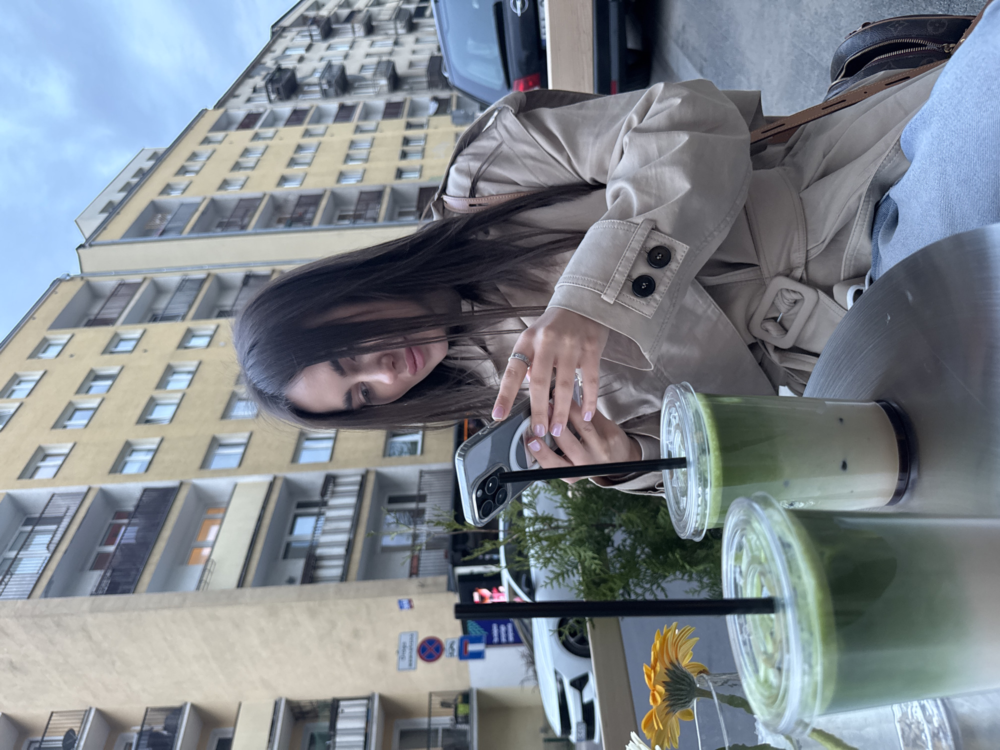A woman is sitting at an outdoor table, holding a smartphone, presumably taking a photo. Two plastic cups with green drinks and black straws are visible on the table. A single yellow flower is in a vase near the drinks. The woman is wearing a tan trench coat. The background features a large yellow building with numerous windows and balconies. Street signs and parked cars are also visible. The location is Warsaw, Poland, based on the architecture and street signs.