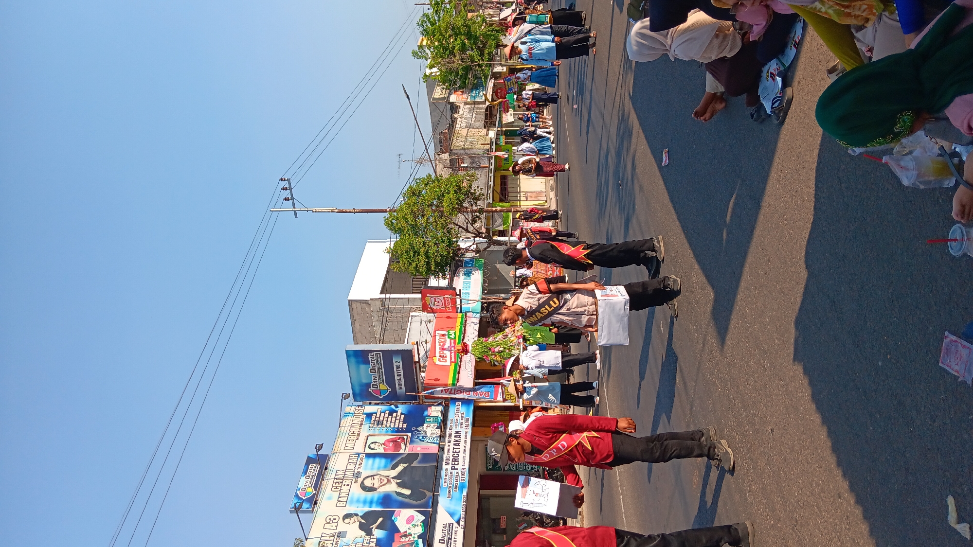 This is a vertical, eye-level, medium shot captured on a sunny day in Tanjung Anom, Indonesia. The image depicts a street scene, likely a parade or procession. In the foreground, several individuals are walking on the asphalt road, dressed in red and black attire with gold sashes. One person holds a white box with a drawing on it, while another carries a similar white box. Behind them, a group of people in various outfits, some wearing hats, are also present on the street. In the background, buildings line the road, featuring numerous signs, including "DANI DIGITAL" and "GEPROSKIN." Trees are visible along the side of the street, and utility poles with wires traverse the sky. To the right of the road, several people are seated on the ground, observing the event. The overall atmosphere suggests a lively community gathering.