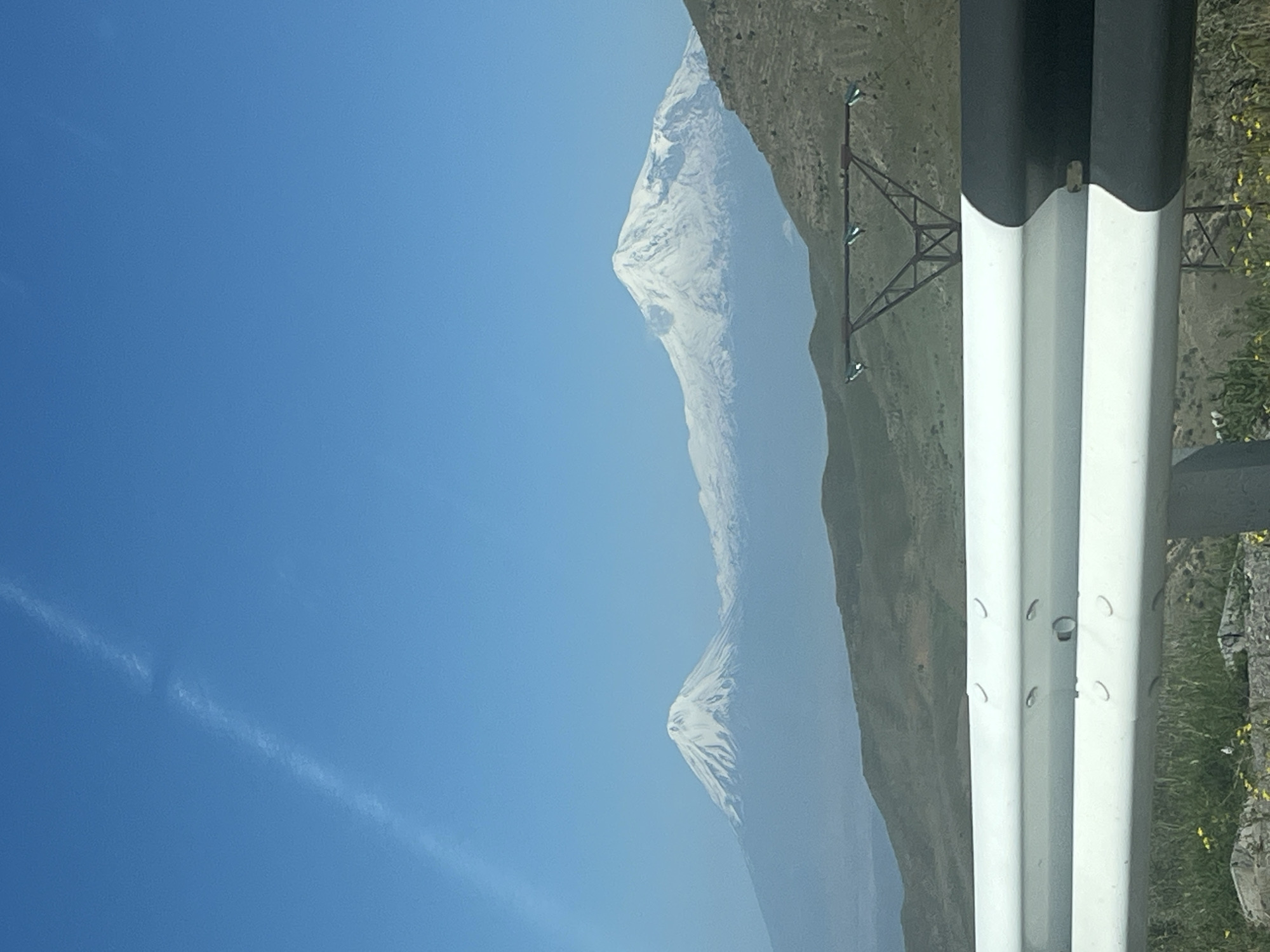 A clear, bright landscape photograph features majestic, snow-capped mountains under a vivid blue sky. The image is oriented vertically, with a prominent black and white corrugated guardrail running along the right side of the frame. The guardrail's white sections have several small, curved indentations and one circular detail.
Behind the guardrail, a green, grassy hillside is visible, dotted with rocks and small yellow flowers. Further into the midground, two large mountains dominate the scene. The closer mountain peak is sharply defined with substantial snow cover. The more distant mountain appears hazier, also displaying snow on its summit. A metal pylon or power line structure stands on the green slope between the guardrail and the mountains.
The sky, a brilliant cerulean blue, fills the left half of the image. A faint, wispy contrail stretches diagonally across the upper left portion of the sky. The lighting suggests a clear day, likely morning or afternoon, with excellent visibility, though atmospheric haze softens the distant features. There is no visible text in the image.