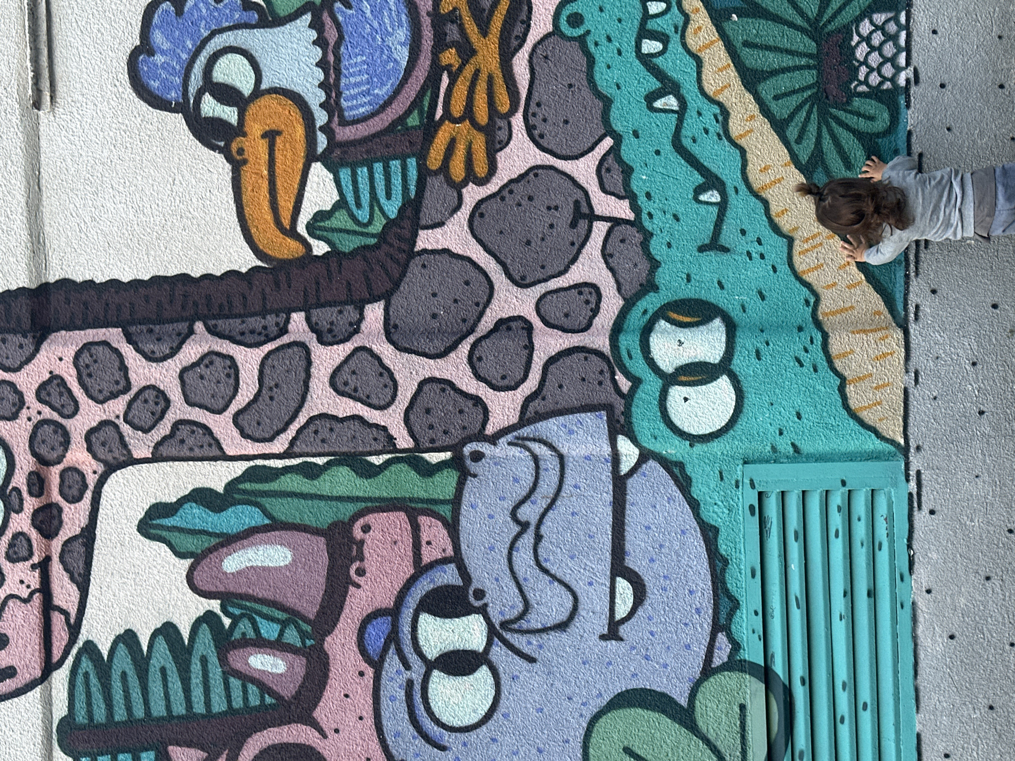 A child is standing in front of a colorful mural on a textured wall, reaching up with both hands. The mural features cartoonish animals, including a giraffe with pink and gray spots, a blue bird with an orange beak, a purple rhinoceros, a light blue hippopotamus-like creature, and a green alligator. The background of the mural includes green foliage and light gray elements. The lower portion of the wall has a turquoise vent. The overall scene suggests an outdoor urban environment in Sofia, Bulgaria.