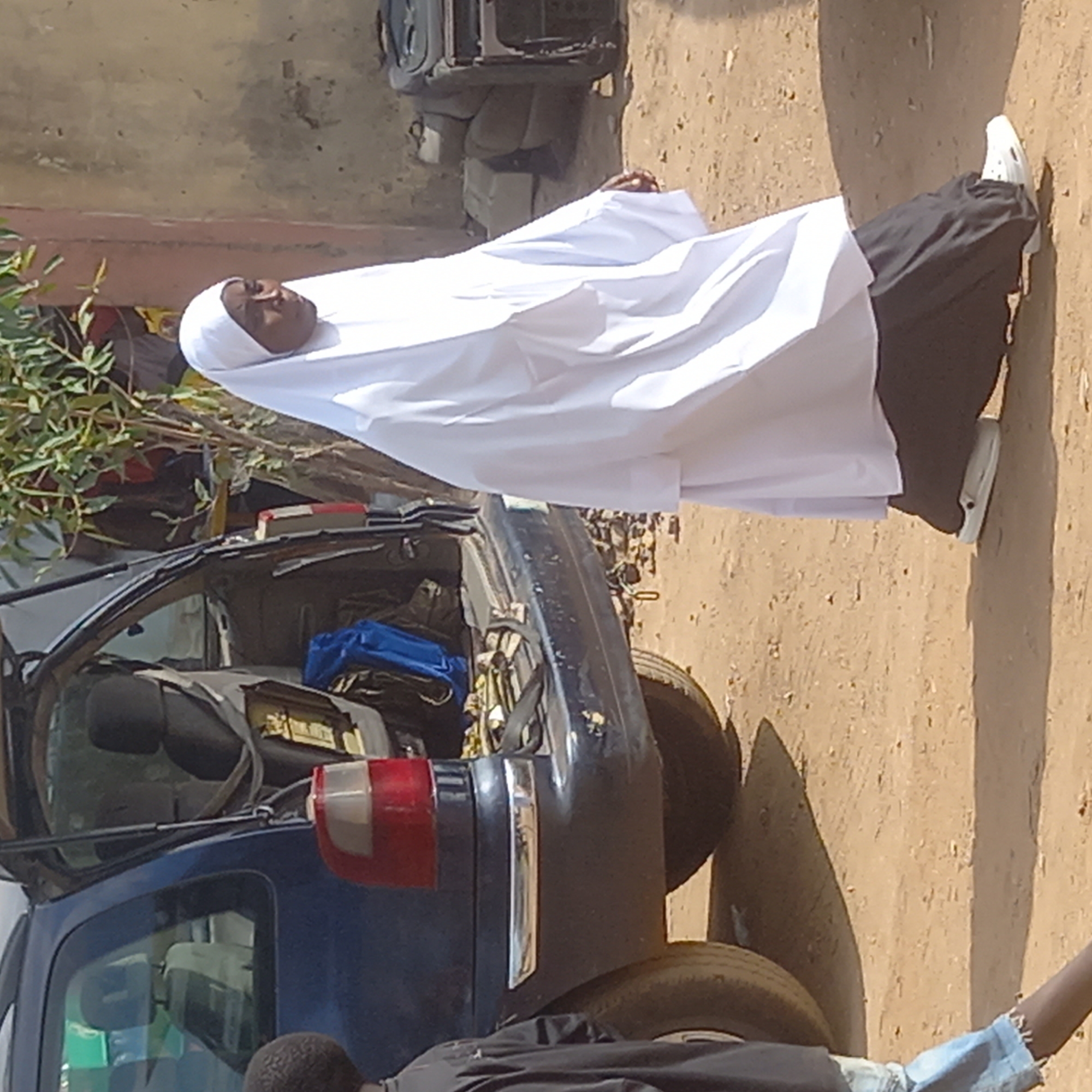 The image shows a woman in a white hijab and black clothing lying near the wreckage of a blue vehicle. The location appears to be outdoors in Dadi Riba, Nigeria, with a dirt ground surface and a building in the background. Another person is partly visible at the bottom of the image.