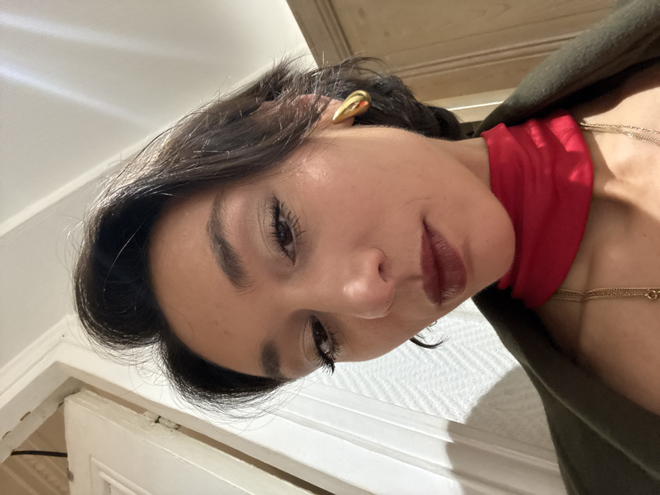 The image displays a low-angle, close-up portrait of an adult female looking directly downwards towards the camera. She has dark hair styled with volume at the crown, a medium skin tone, and wears dark red lipstick with visible eye makeup, including mascara and light eyeshadow. A single gold-toned, teardrop-shaped earring is visible on her right earlobe. She is dressed in a red high-neck top, possibly a turtleneck, beneath an olive green or dark green outer garment. Multiple delicate gold-toned chain necklaces are worn around her neck. The background depicts an indoor environment with a white doorframe on the left and a wooden panel or piece of furniture on the right. Light reflections are visible on the upper right side, indicative of an overhead light source. The specific location within Paris, France, cannot be determined from the visual evidence presented in the image.